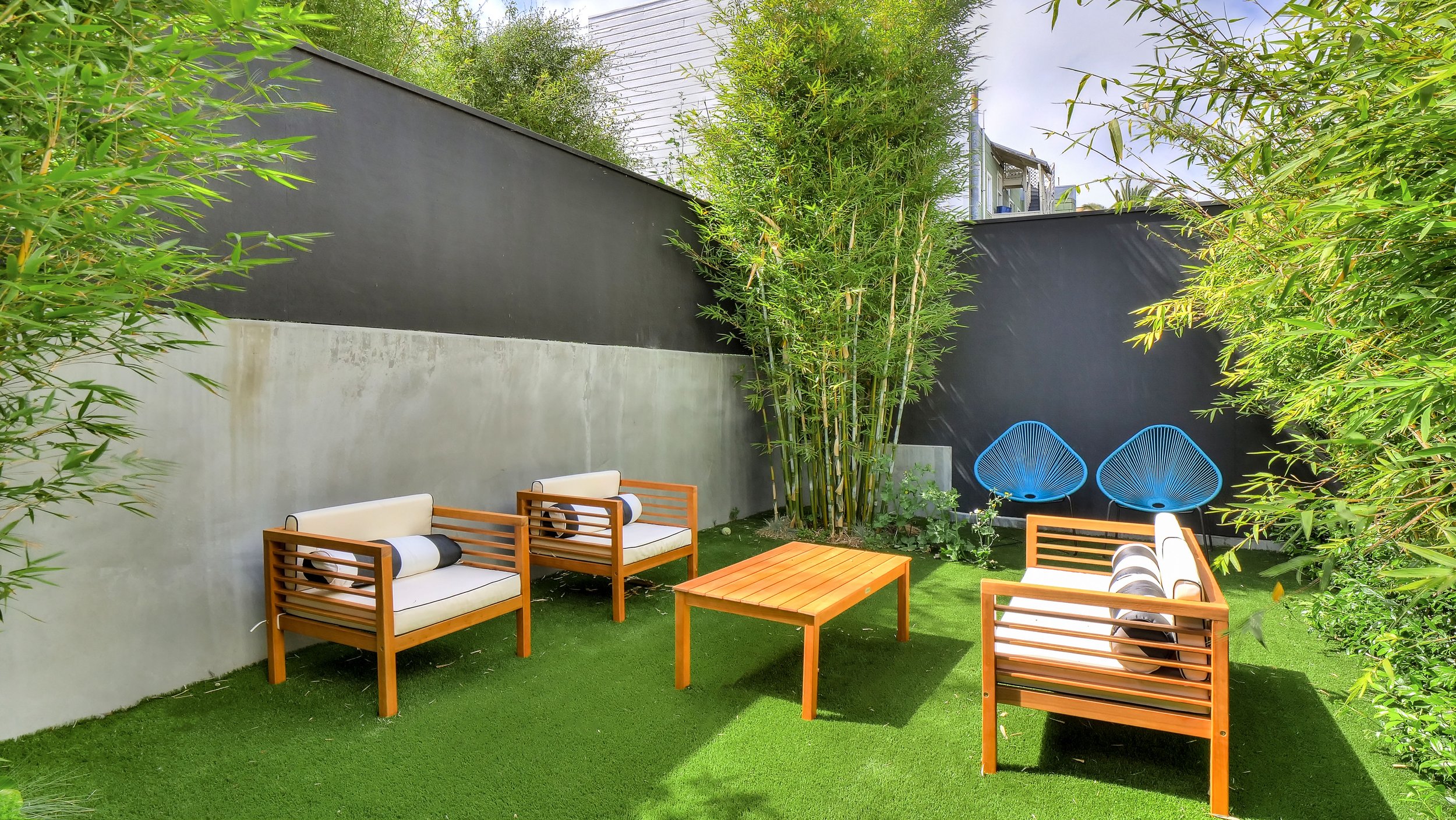 Small backyard with artificial grass, surrounded by high walls with plants. There are three wooden benches with white cushions and black pillows, a matching wooden coffee table, and two blue chairs with circular design.