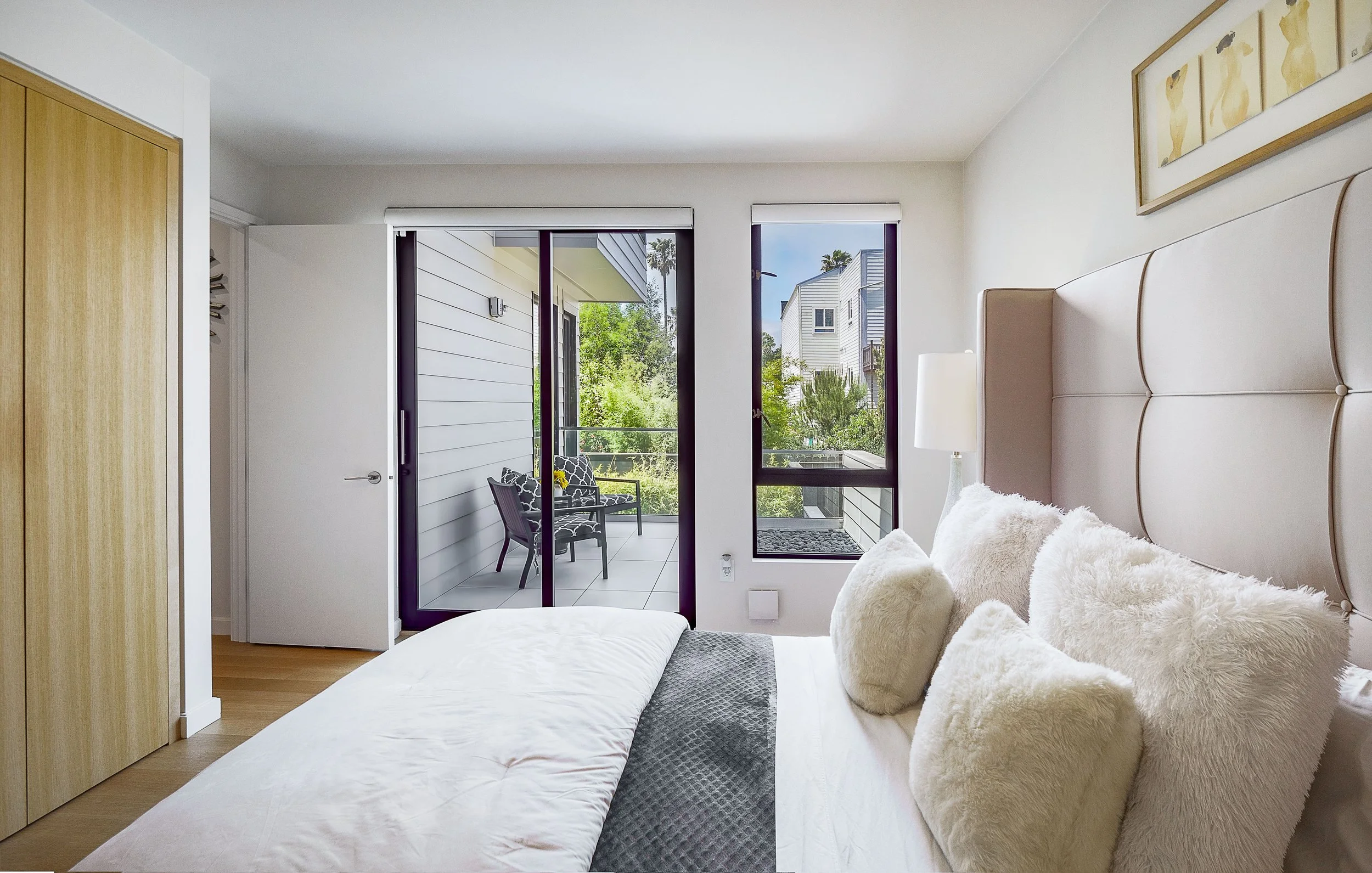 Bedroom with bed, plush pillows, nightstand with lamp, sliding glass door leading to balcony with outdoor seating, and cityscape view.