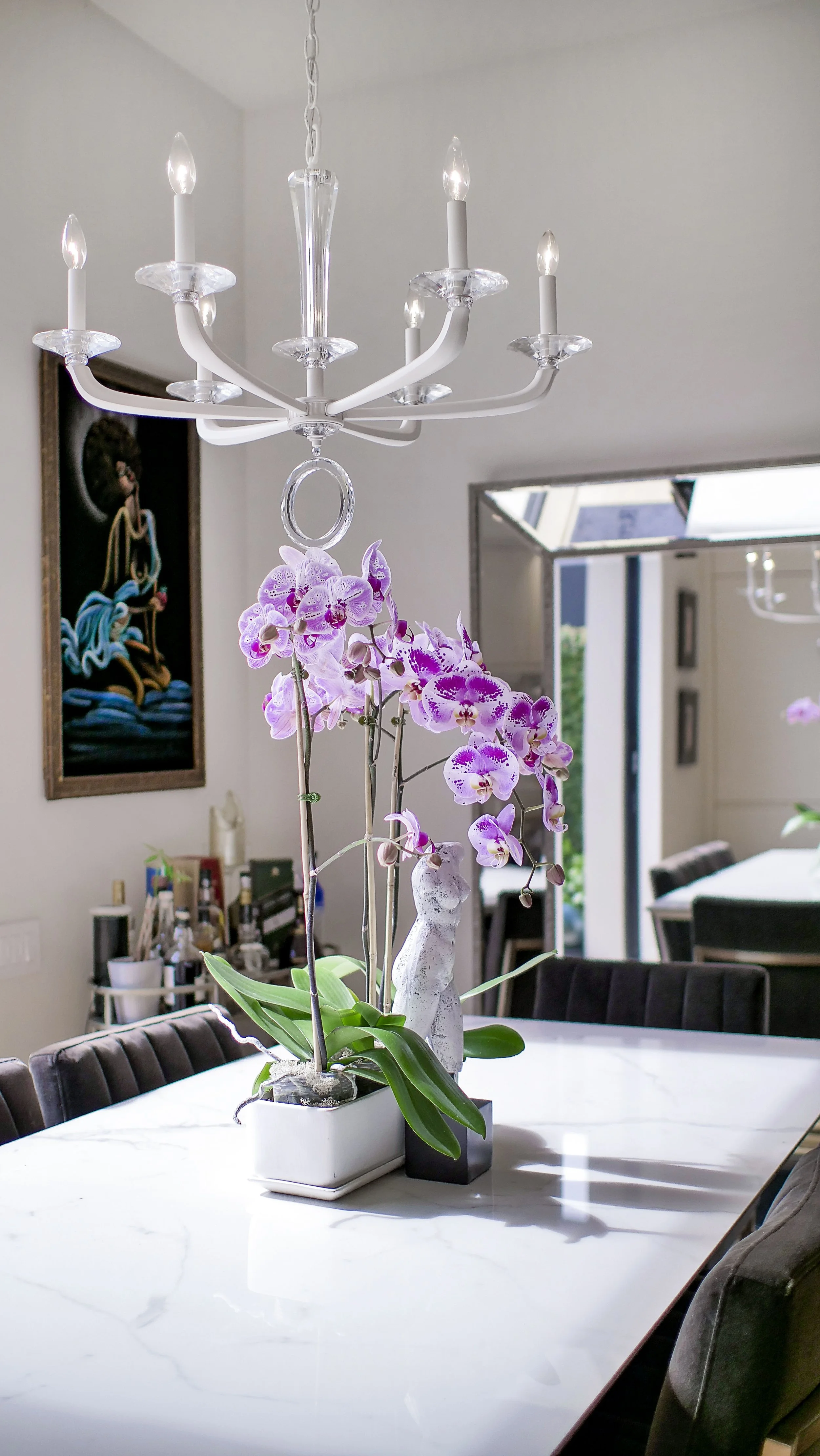 A dining room with a white marble table, decorated with pink orchids in ceramic and black pots. A white chandelier hangs above the table, and a painting is on the wall. There is a window in the background letting in natural light.