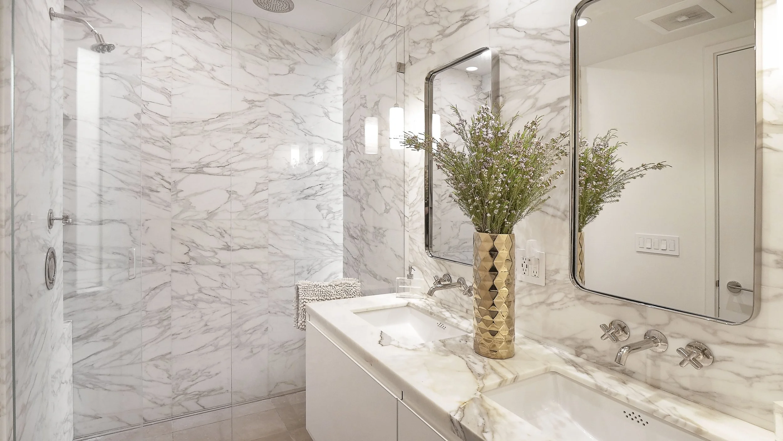 A modern bathroom with marble walls, a double sink vanity with marble countertop, two large mirrors, and a decorative vase with purple flowers. There is also a walk-in shower with a rain showerhead.