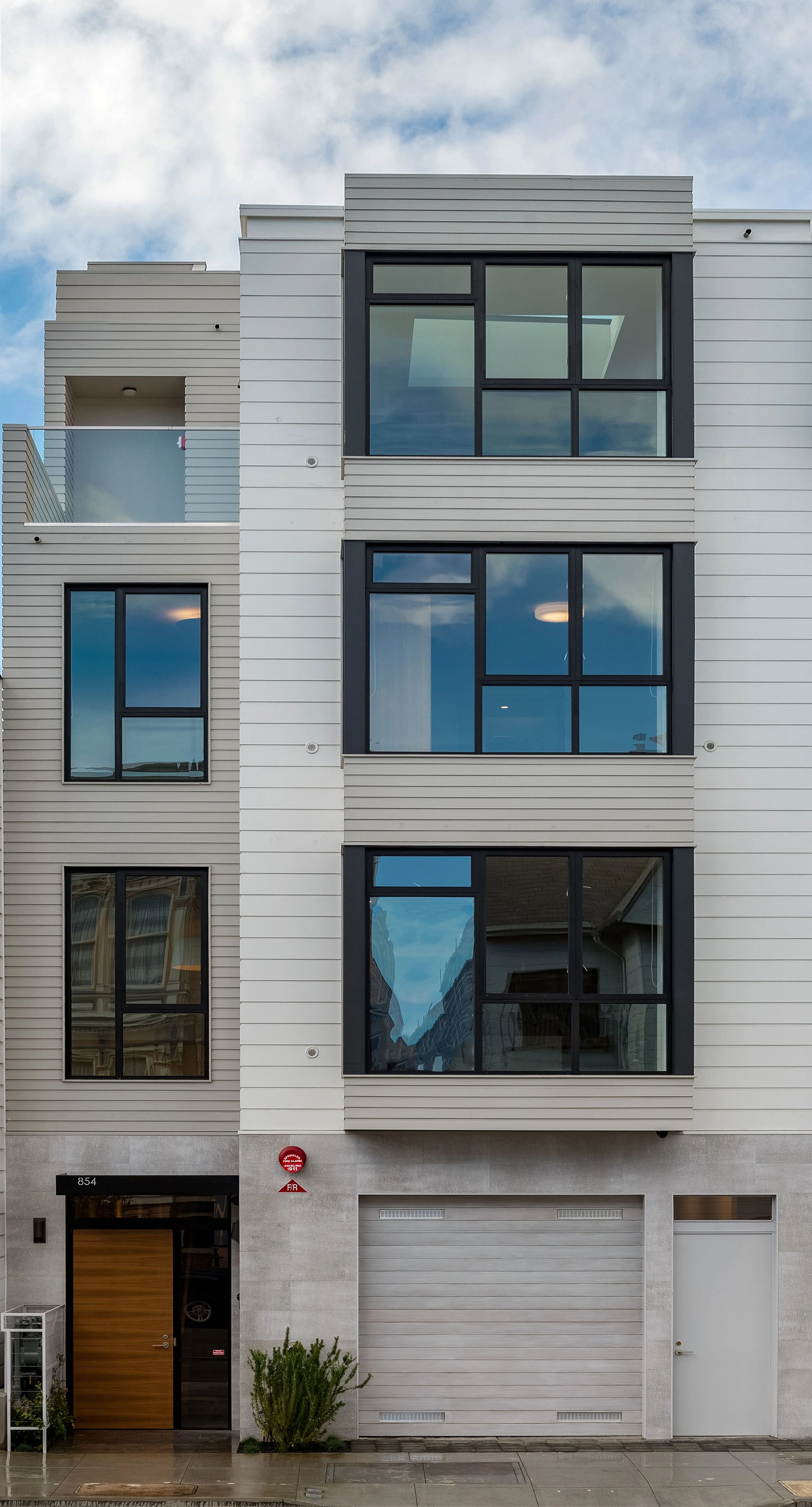 Modern multi-story building with large glass windows and a garage, small plants at entrance, partly cloudy sky.