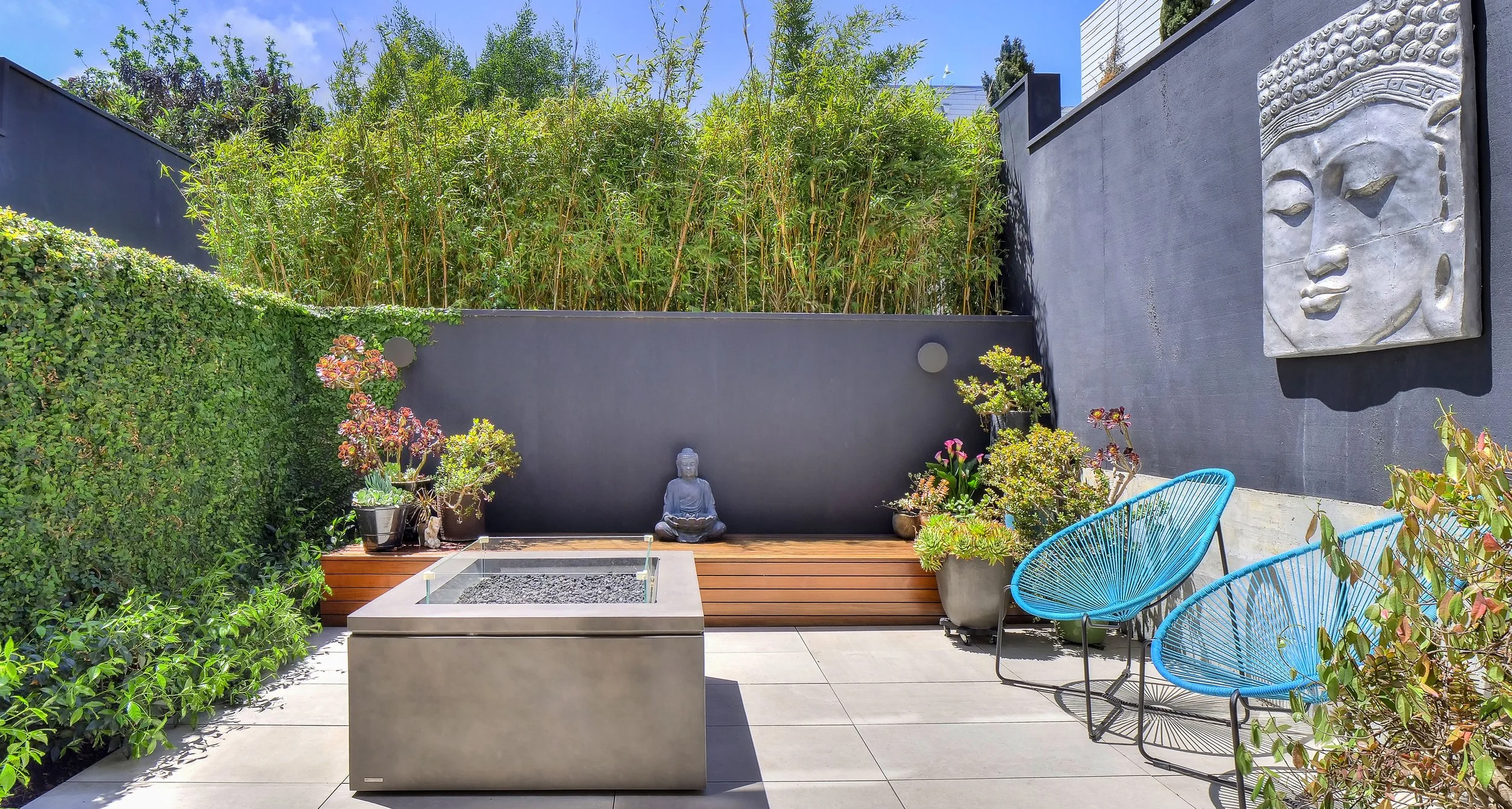 Modern outdoor patio with potted plants, a Buddha statue, enclosed by greenery and walls, with blue circular chairs and a fire pit.