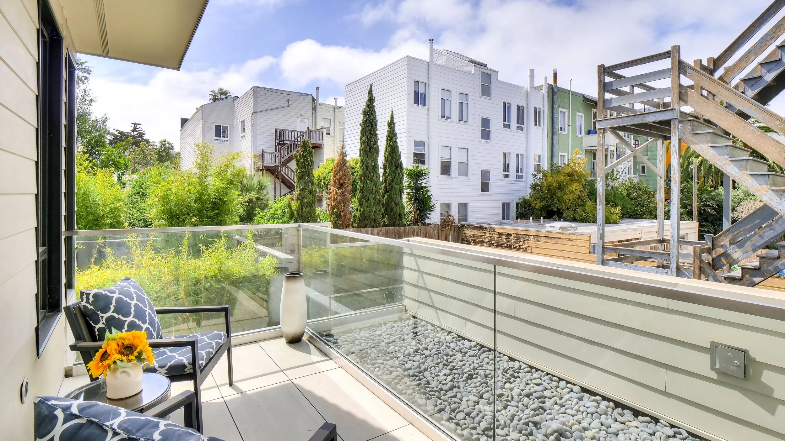 Balcony with two black and white patterned chairs, a small table with a sunflower in a white vase, and a tall white vase, overlooking a garden with greenery and trees, with residential buildings in the background and an outdoor staircase on the right