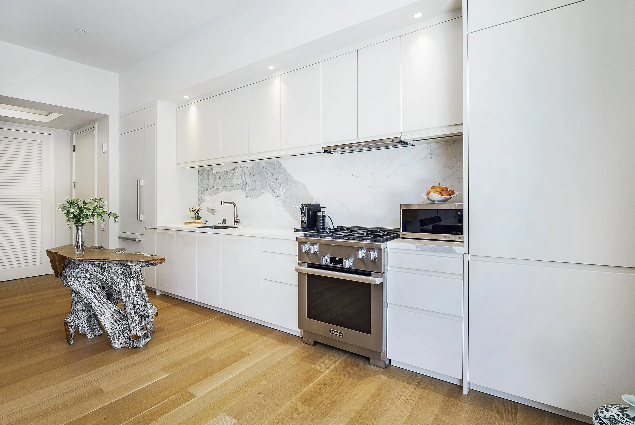 Modern kitchen with white cabinets, marble backsplash, stainless steel stove, microwave, and coffee maker, wood flooring, and a small wooden table with a plant.