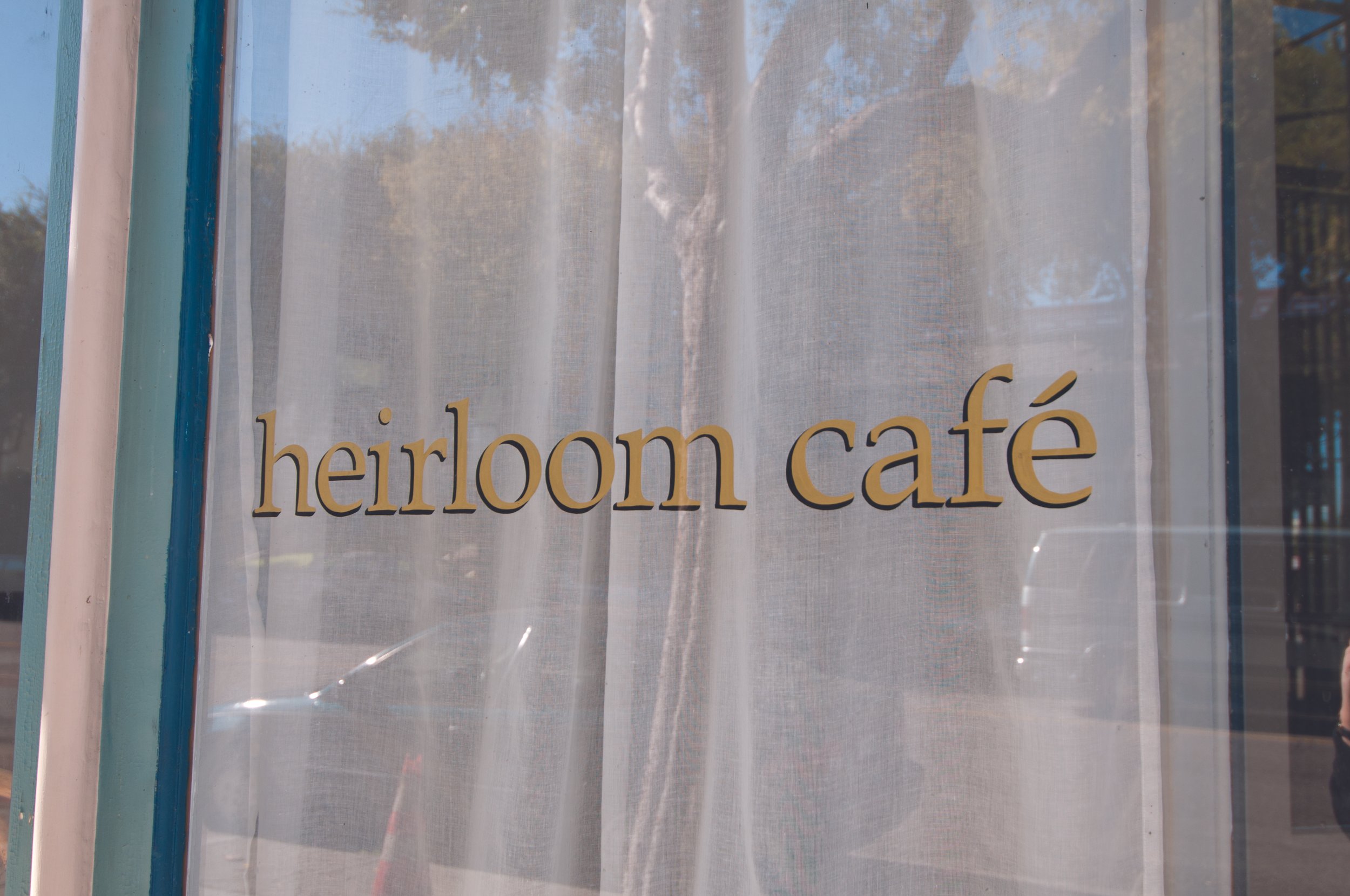 Sign reading 'heirloom café' on a window with a white curtain behind it and reflections of trees and buildings outside.