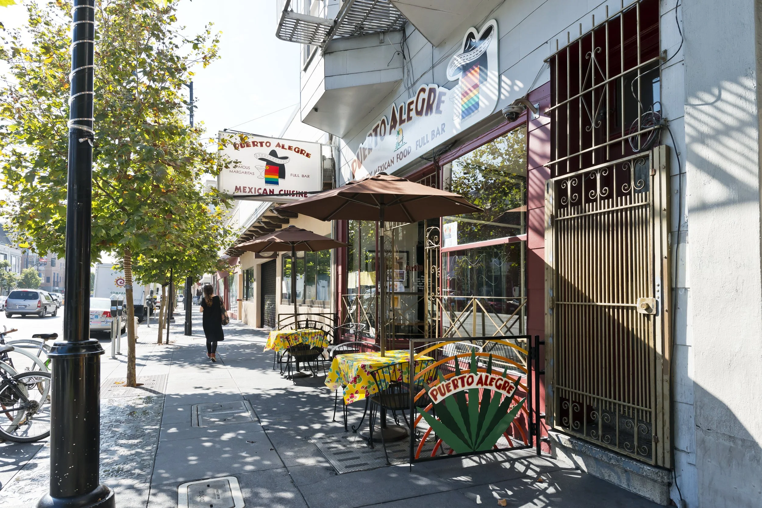 Street view of Puerto Alegre Mexican restaurant with outdoor seating, tables with colorful tablecloths, umbrellas, and trees lining the sidewalk.