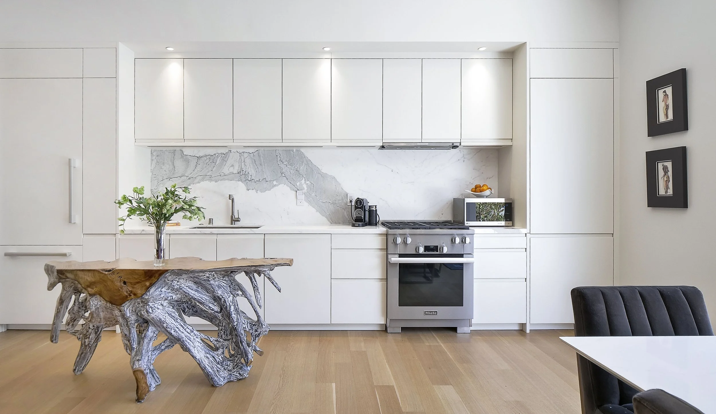 Modern white kitchen with marble backsplash, wooden floor, and a rustic wooden dining table with a plant on top.