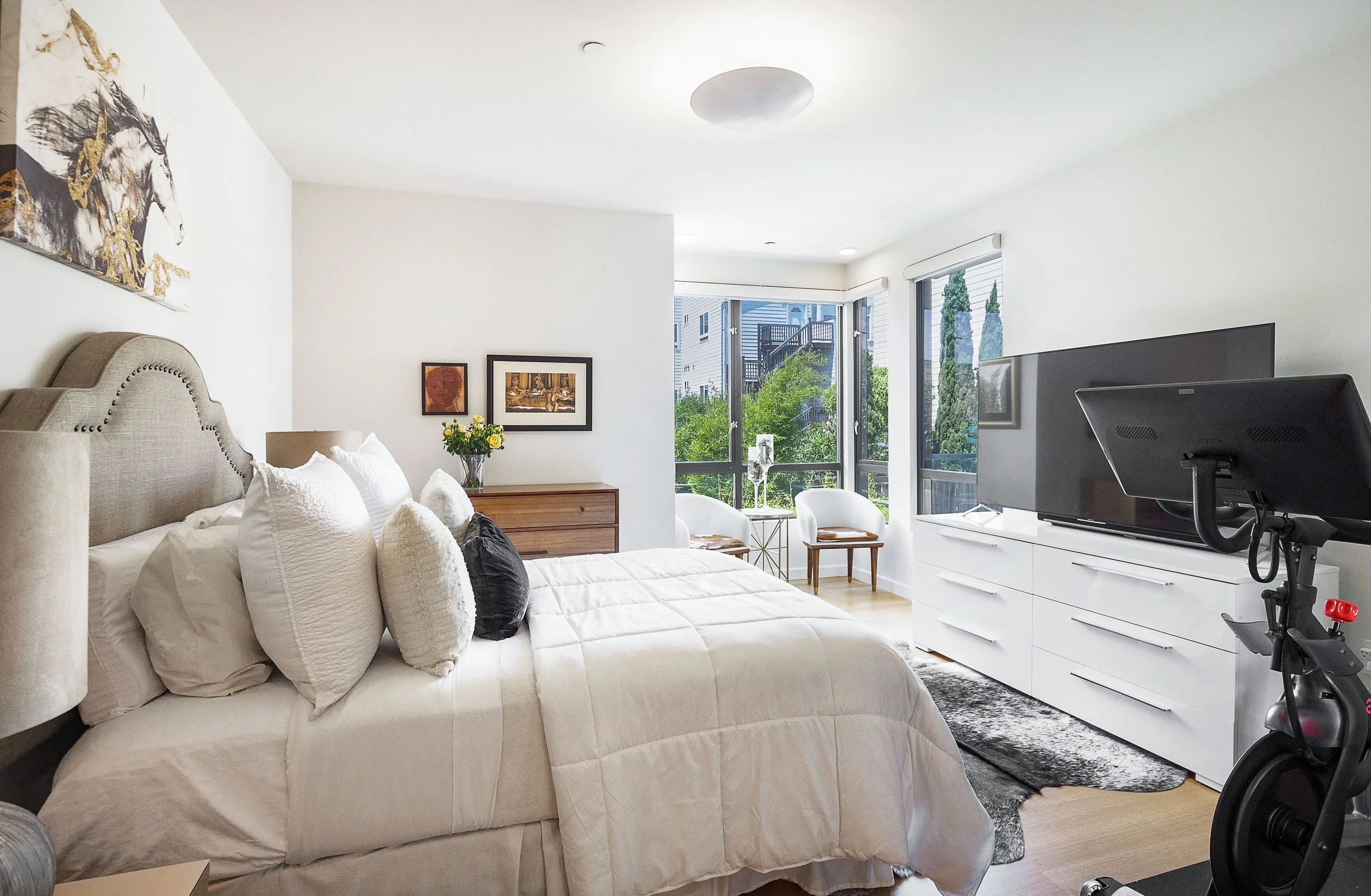 Bright and modern bedroom with a large bed, white bedding, multiple pillows, a wooden dresser, and a TV on a white dresser. Large windows with outdoor greenery and a balcony, with a stationary exercise bike in the corner.