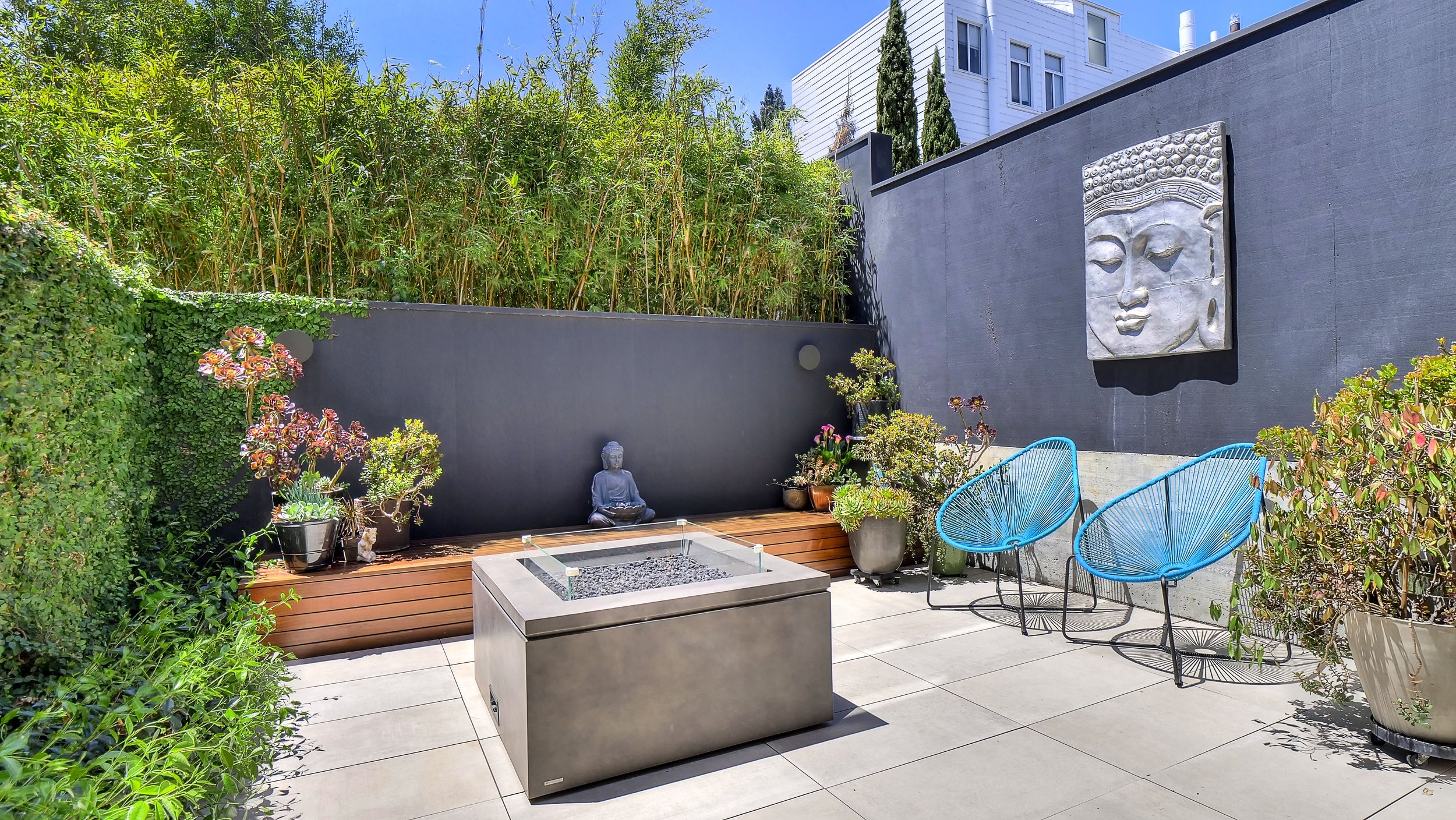 Modern outdoor patio with potted plants, a Buddha statue, a fire feature, blue chairs, and a large decorative Buddha face wall sculpture, surrounded by greenery and tall bamboo.