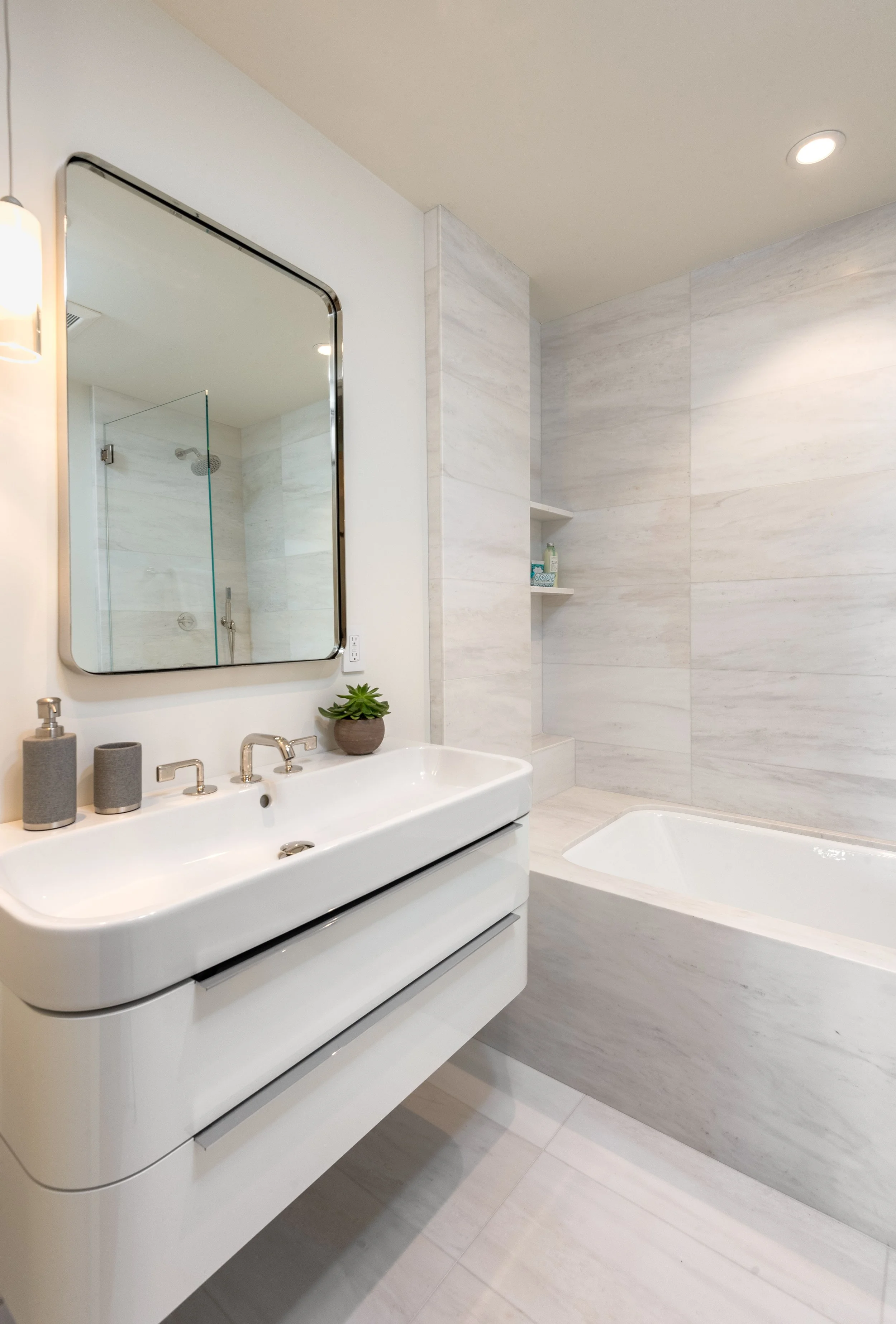 Modern bathroom with a white vanity, a rectangular mirror, a small potted plant, and a bathtub with marble surround.