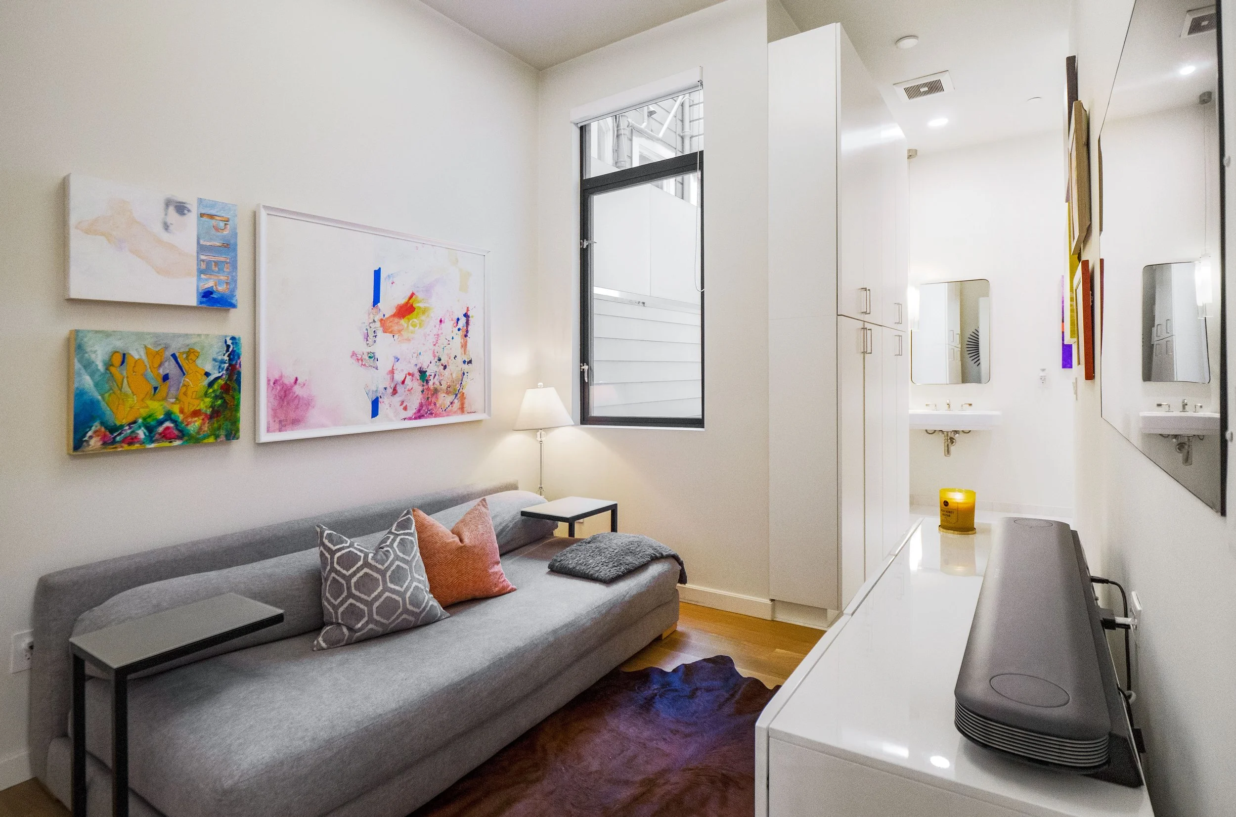 A cozy living space with a gray sofa adorned with patterned and solid pillows, a small side table, a white dresser with a black speaker, and framed abstract artwork on white walls. A lamp sits beside a window with black framing, and a partial view of