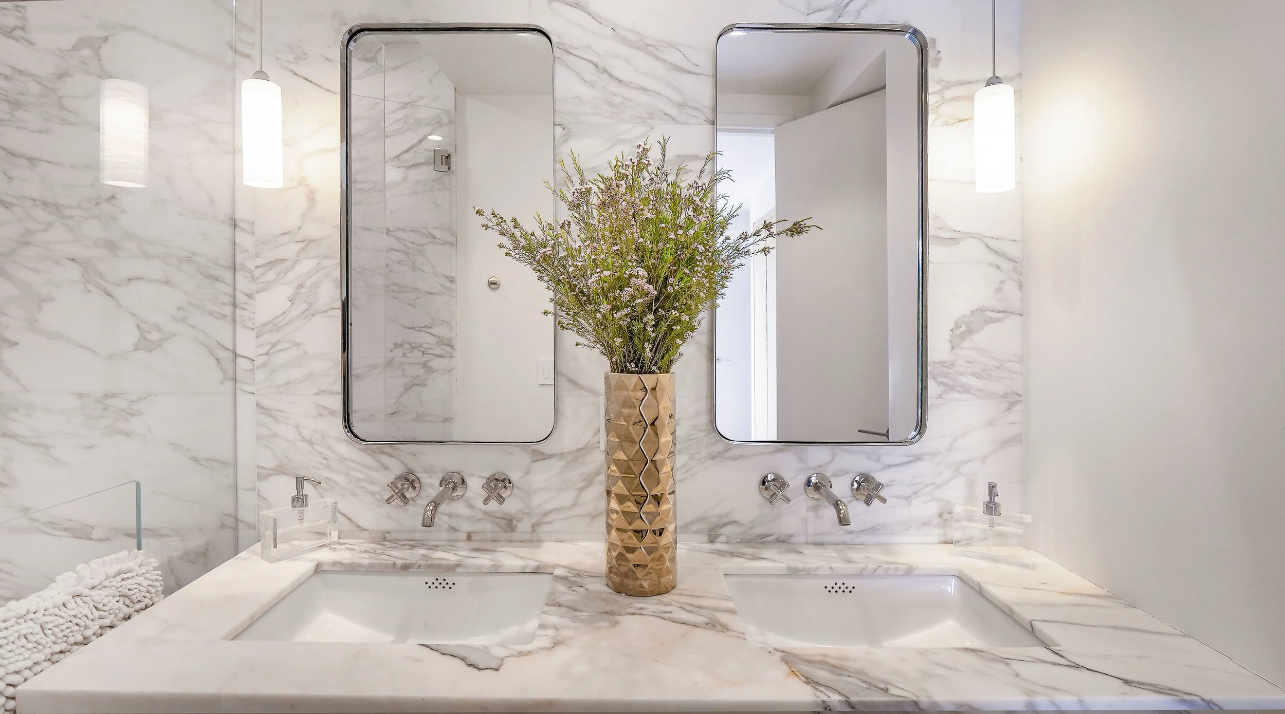 Double bathroom vanity with marble countertop, two rectangular mirrors, wall-mounted faucets, a tall vase with a large flower arrangement in the center, marble wall, and two cylindrical hanging lights.