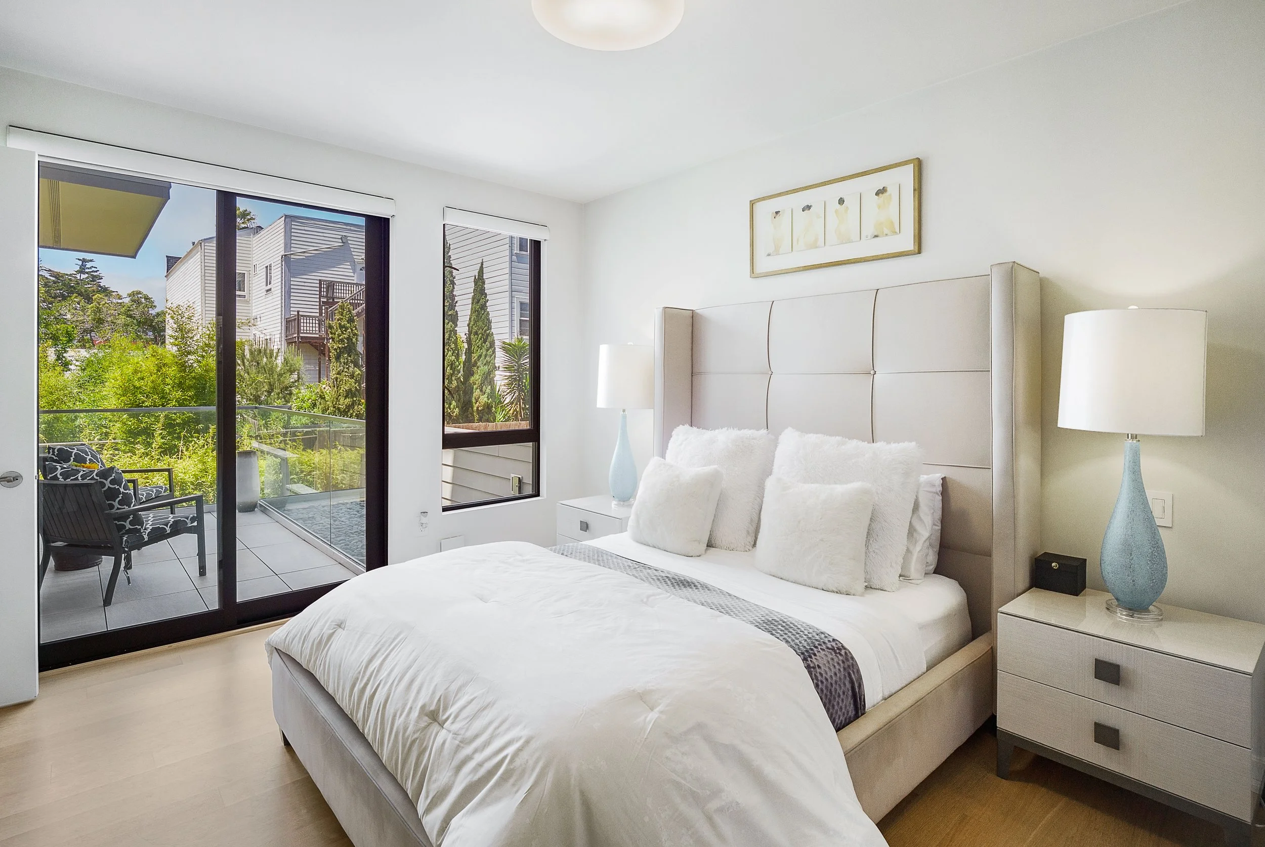 Bright bedroom with a large bed, white bedding, and multiple white pillows, next to sliding glass doors leading to a small balcony with outdoor seating, palm trees, and neighboring houses visible outside.