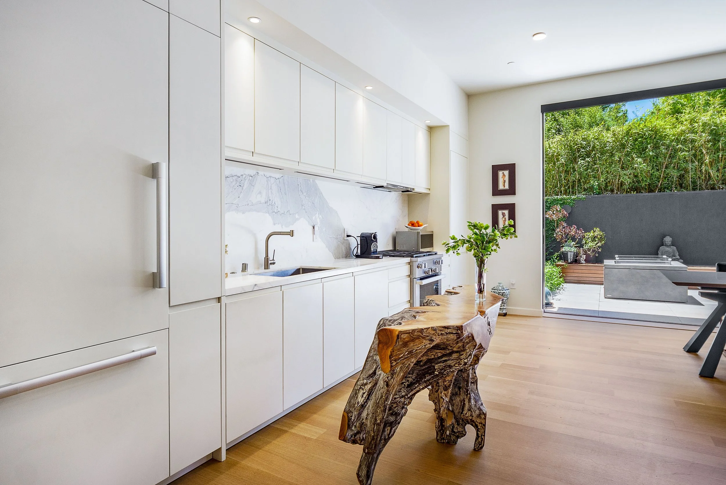 Modern kitchen with white cabinets, marble backsplash, wooden floor, and a rustic wooden table with a glass vase of green plants. Sliding glass door opens to an outdoor patio with outdoor furniture and greenery.