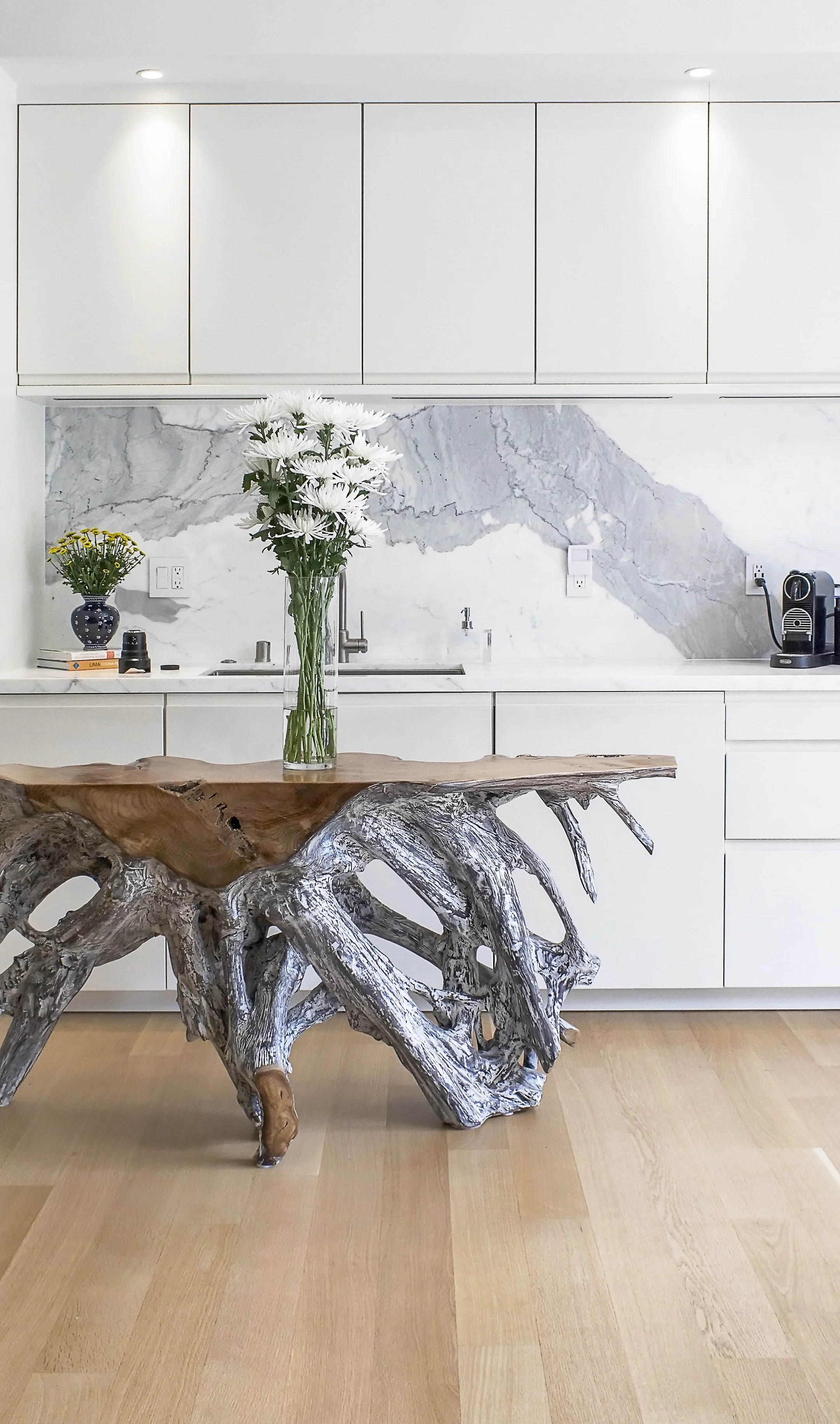 Modern kitchen with white cabinets, marble backsplash, a wooden table with driftwood base, and a glass vase with white flowers.