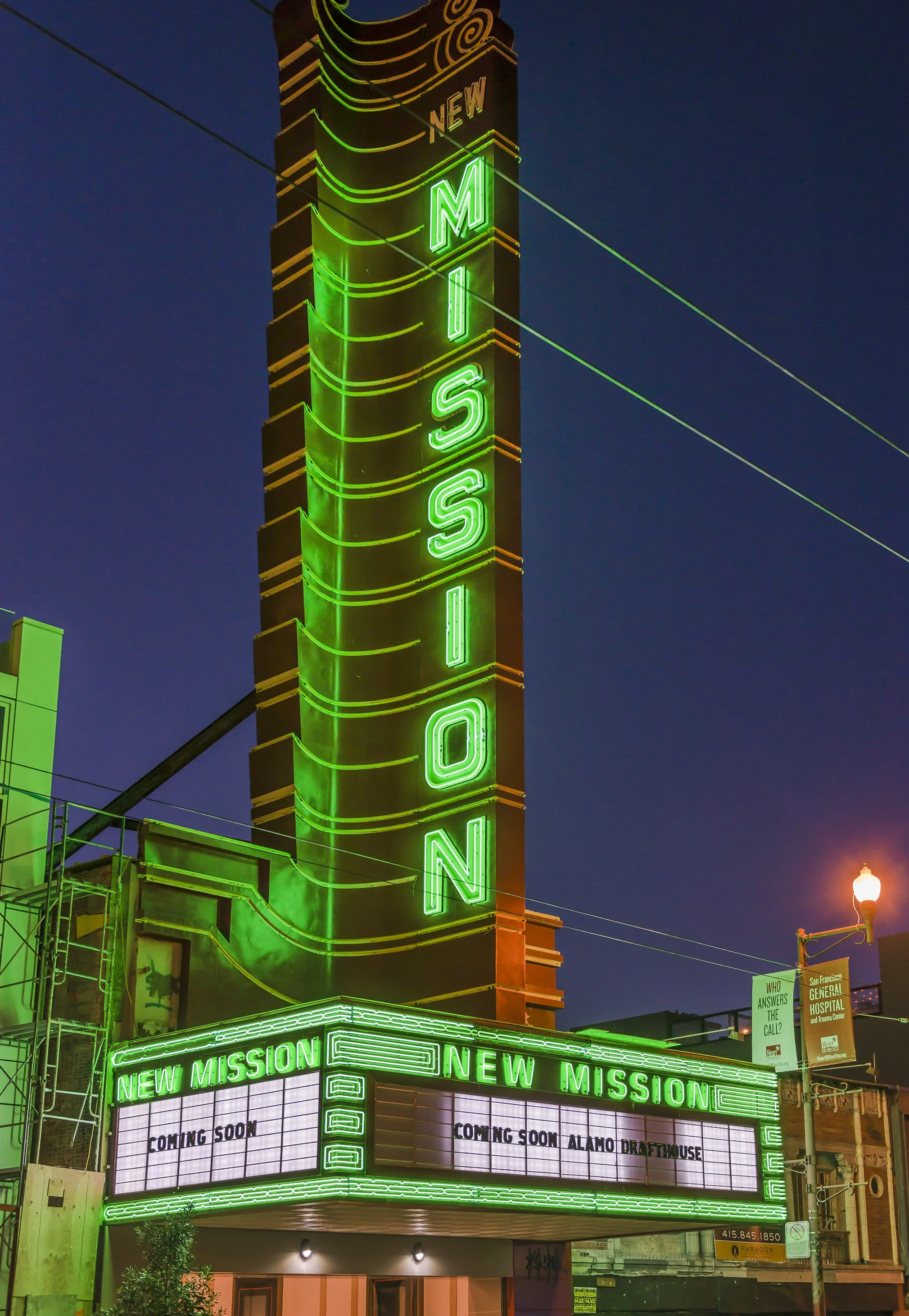 Night view of a neon illuminated sign reading 'MISSION' vertically on a tall tower, with a marquee below displaying updates for a new mission at Alamo Draft House in green neon lights.
