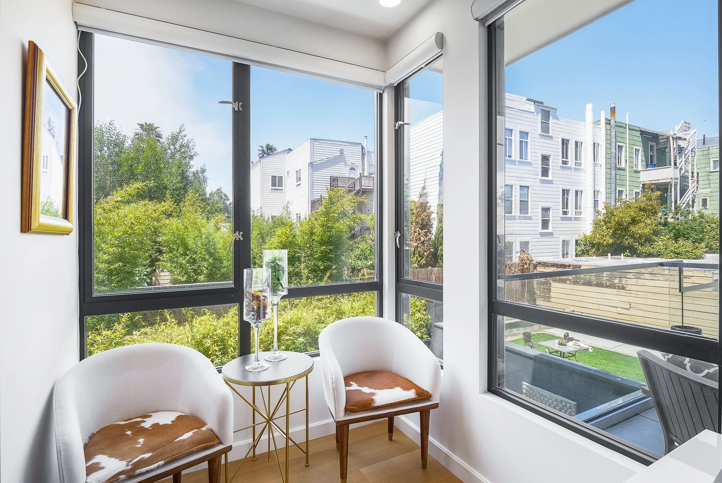 Sunroom with large windows, white walls, two white chairs with cowhide cushions, a gold side table with glass vases, trees and colorful buildings outside.