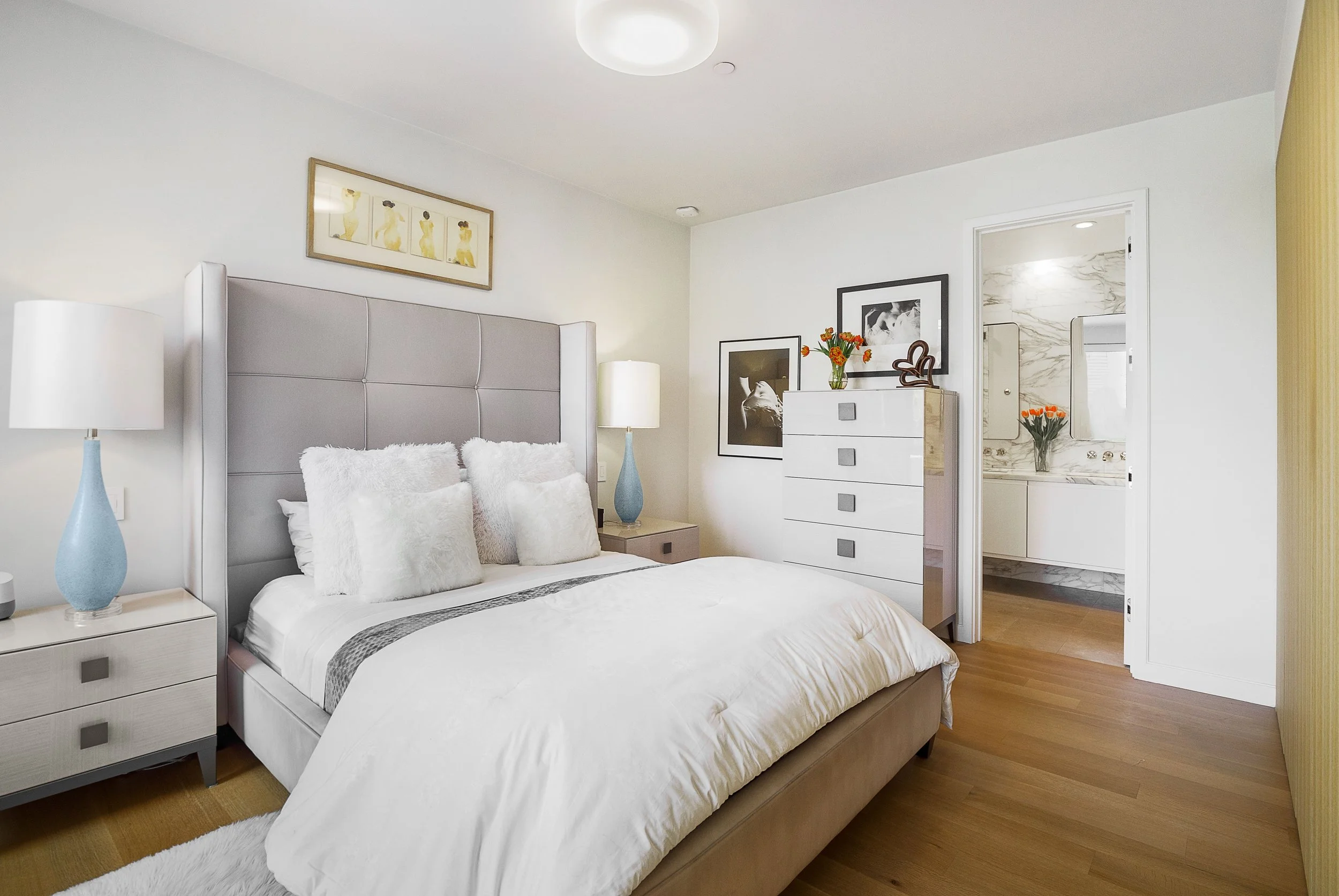 A modern bedroom with a large bed, white bedding, plush pillows, bedside lamps, framed artwork, and a view into a bathroom with a marble vanity and orange flowers.