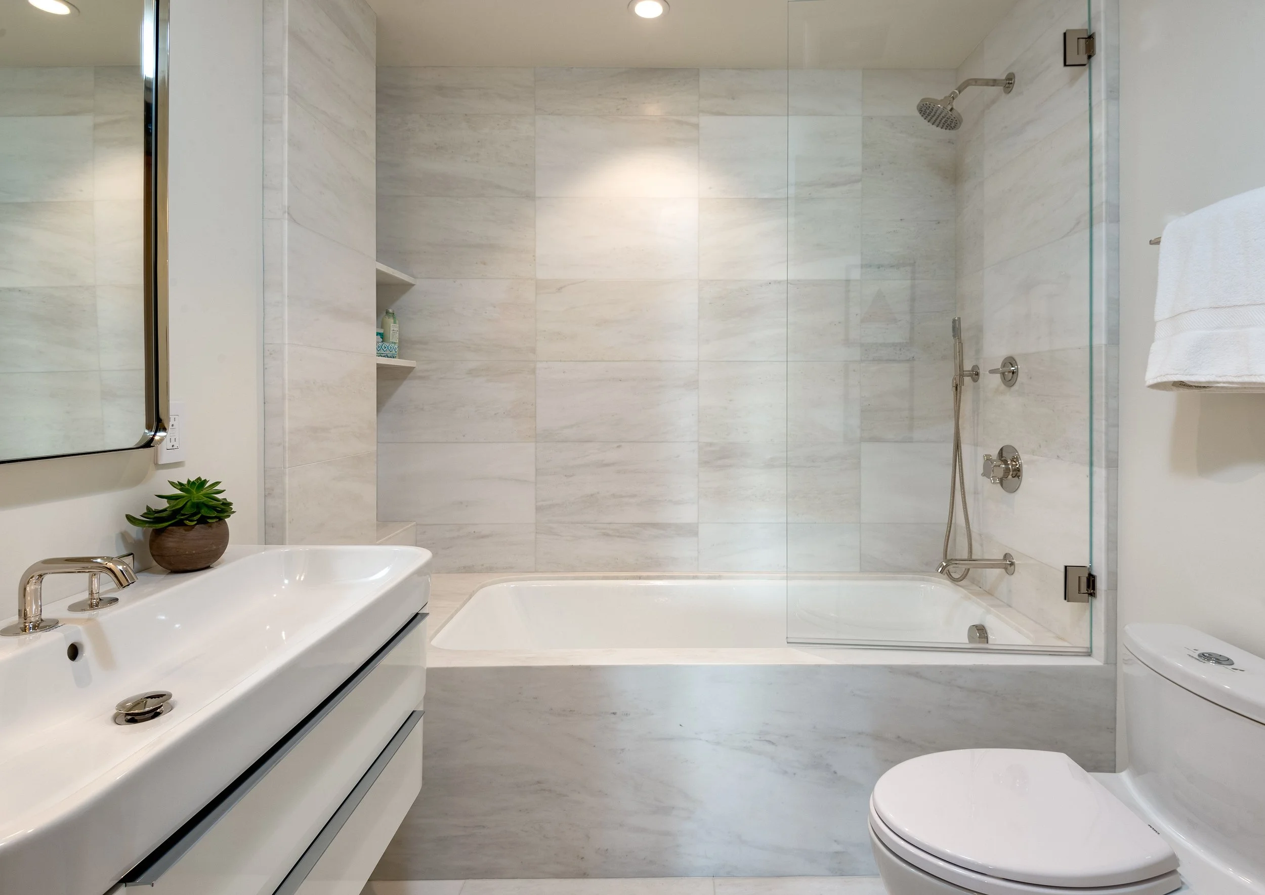 Modern bathroom with white vanity, mirror, small plant, sink, bathtub with glass partition, showerhead, white toilet, and beige marble walls and floor.