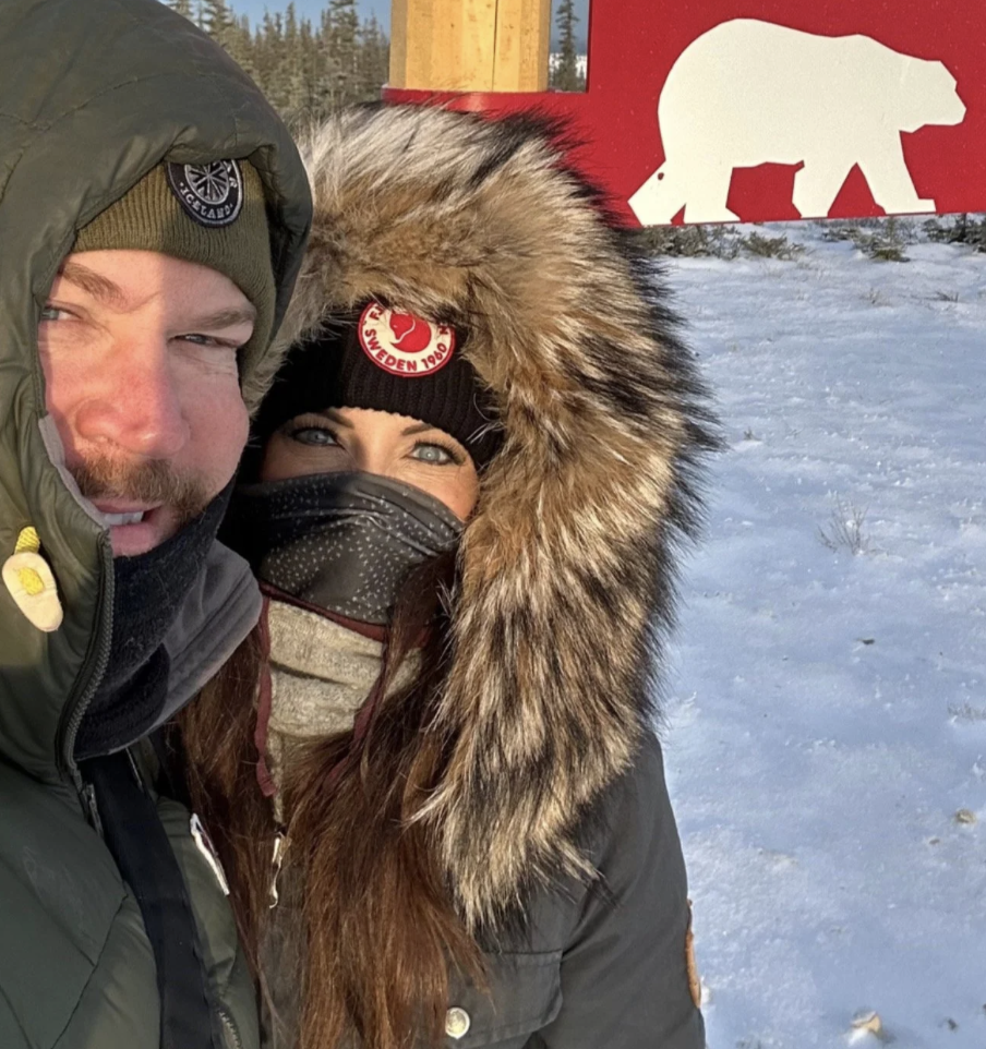 A man and woman in winter clothing taking a selfie outdoors in a snowy area near a red sign with a white bear silhouette. The woman has a fur-lined hood and wears a mask, and the man is wearing a coat and a beanie.