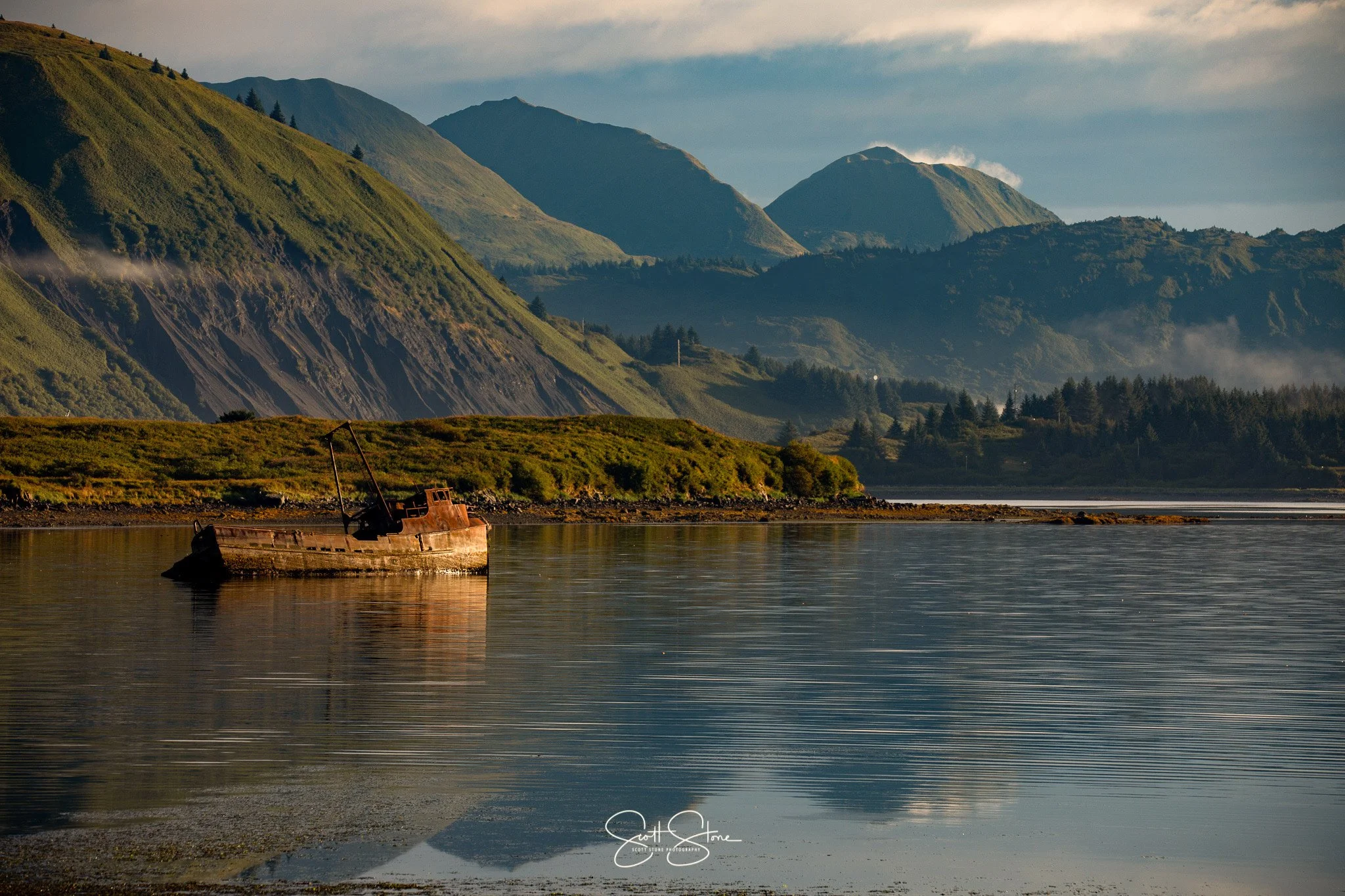 A rusted shipwreck in calm water with green hills and mountains in the background during sunset or sunrise.