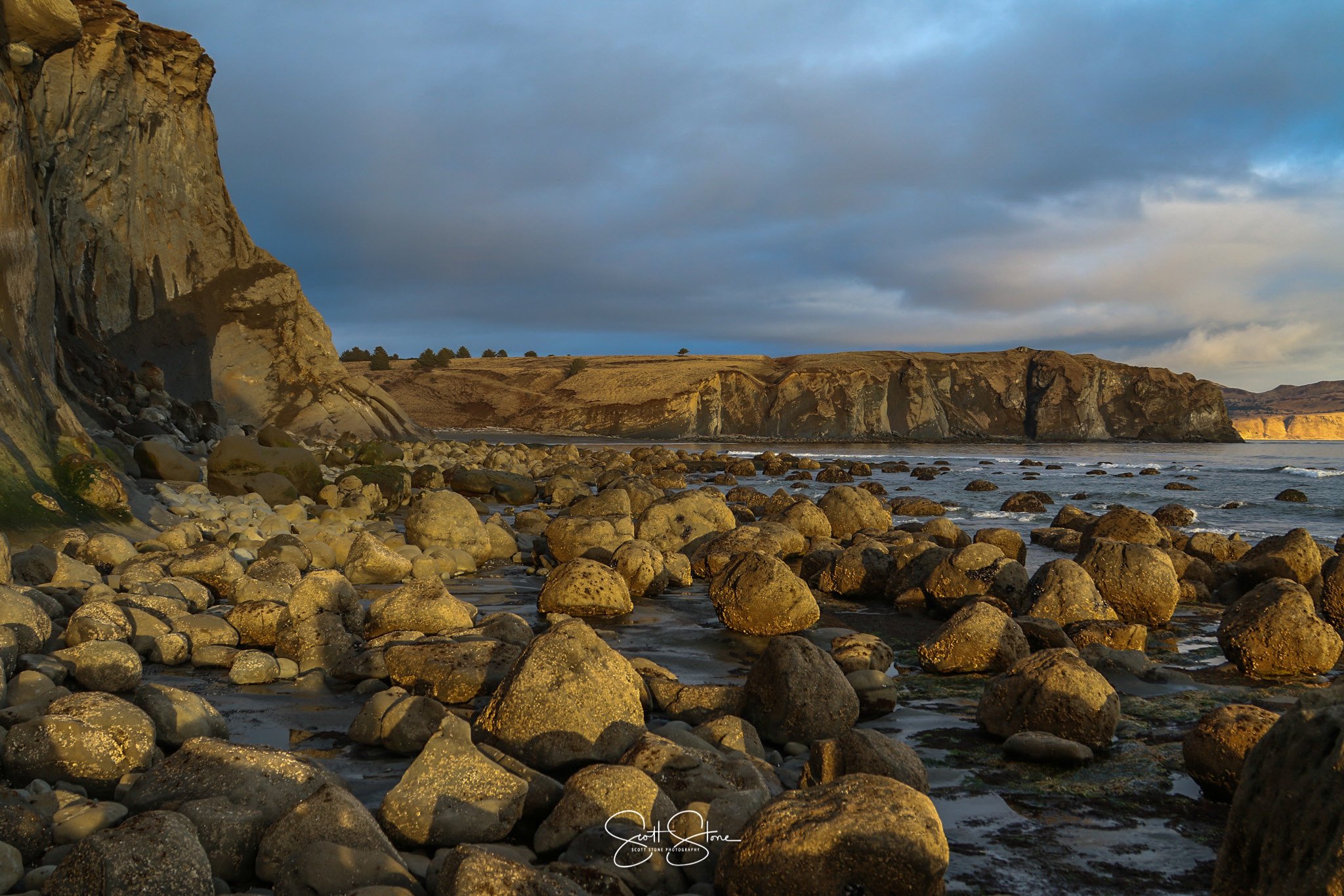 Rocky shoreline with large boulders, cliffs on the left side, and ocean waves, under a partly cloudy sky during sunset.