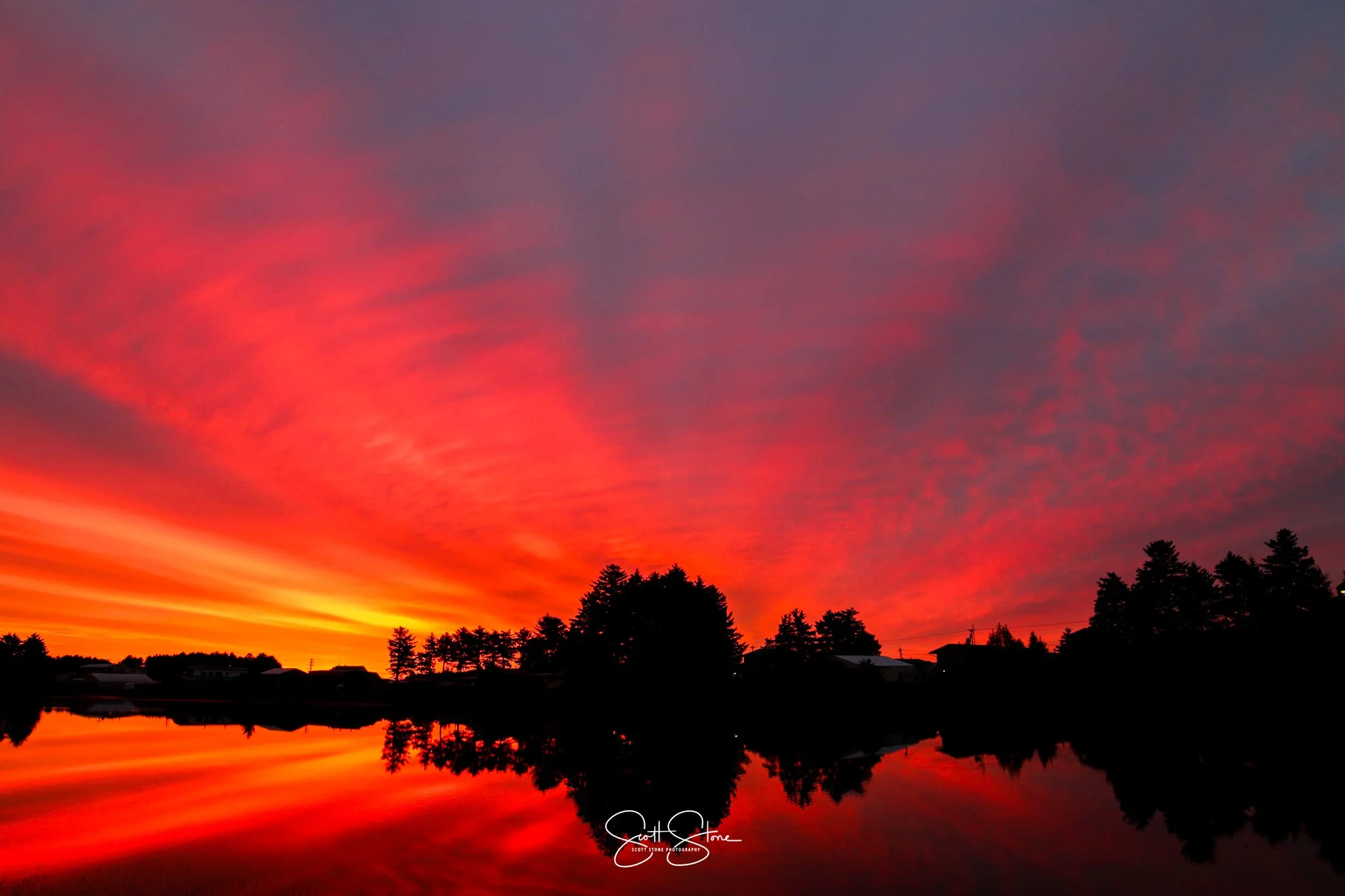 Vivid sunset with orange and pink clouds reflecting on a calm lake, silhouetted trees and houses along the shoreline.