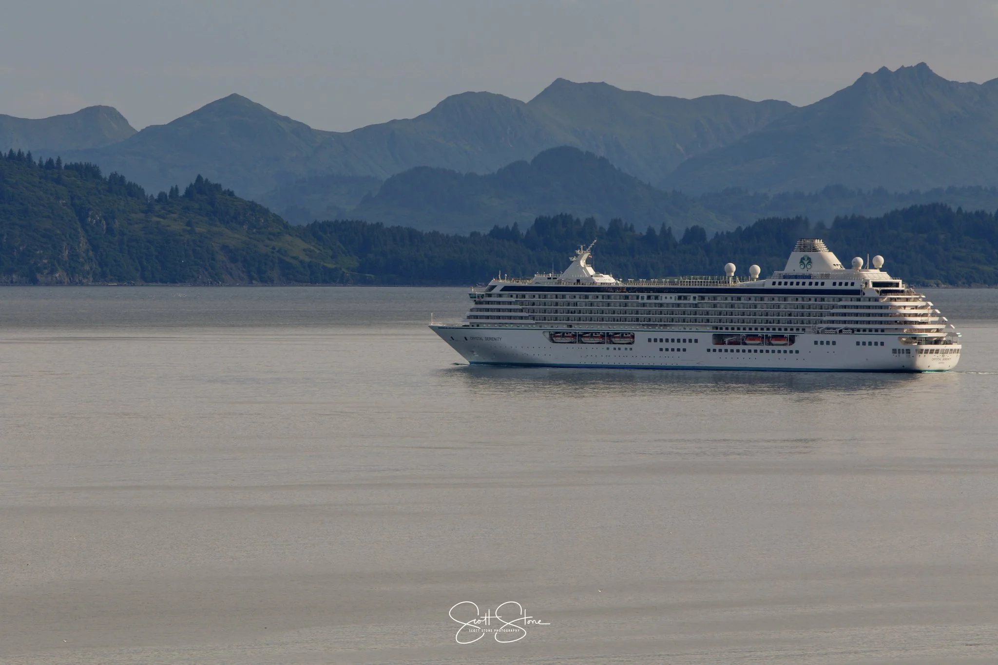 A white cruise ship sailing on a calm body of water with lush green hills and mountains in the background.