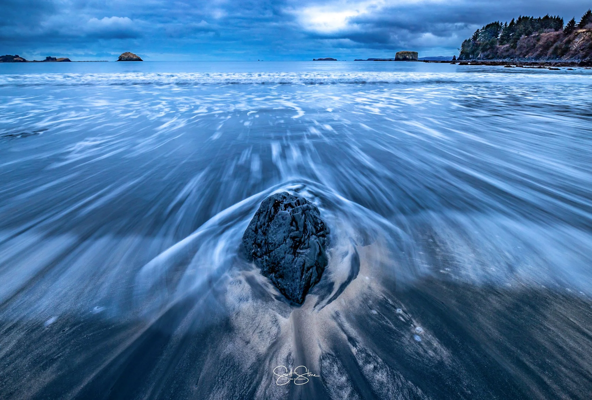 A long exposure photo of a rocky beach with waves crashing around a central large dark rock, and distant islands and cliffs under a cloudy sky.