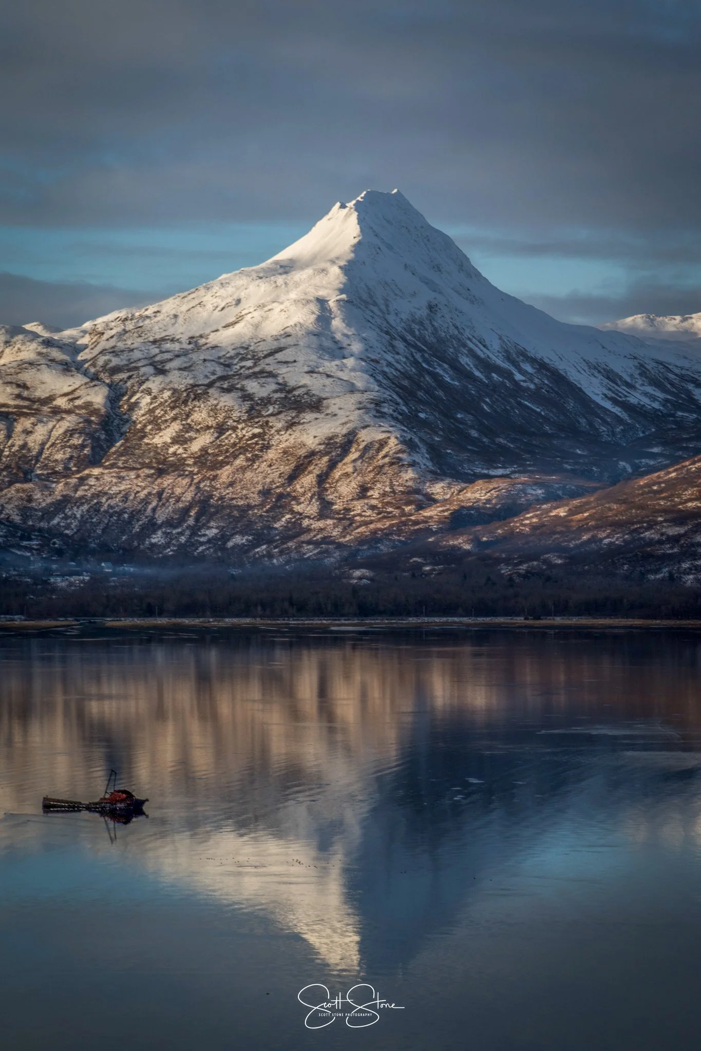 Snow-capped mountain reflected in a calm body of water with a small boat floating in the foreground.