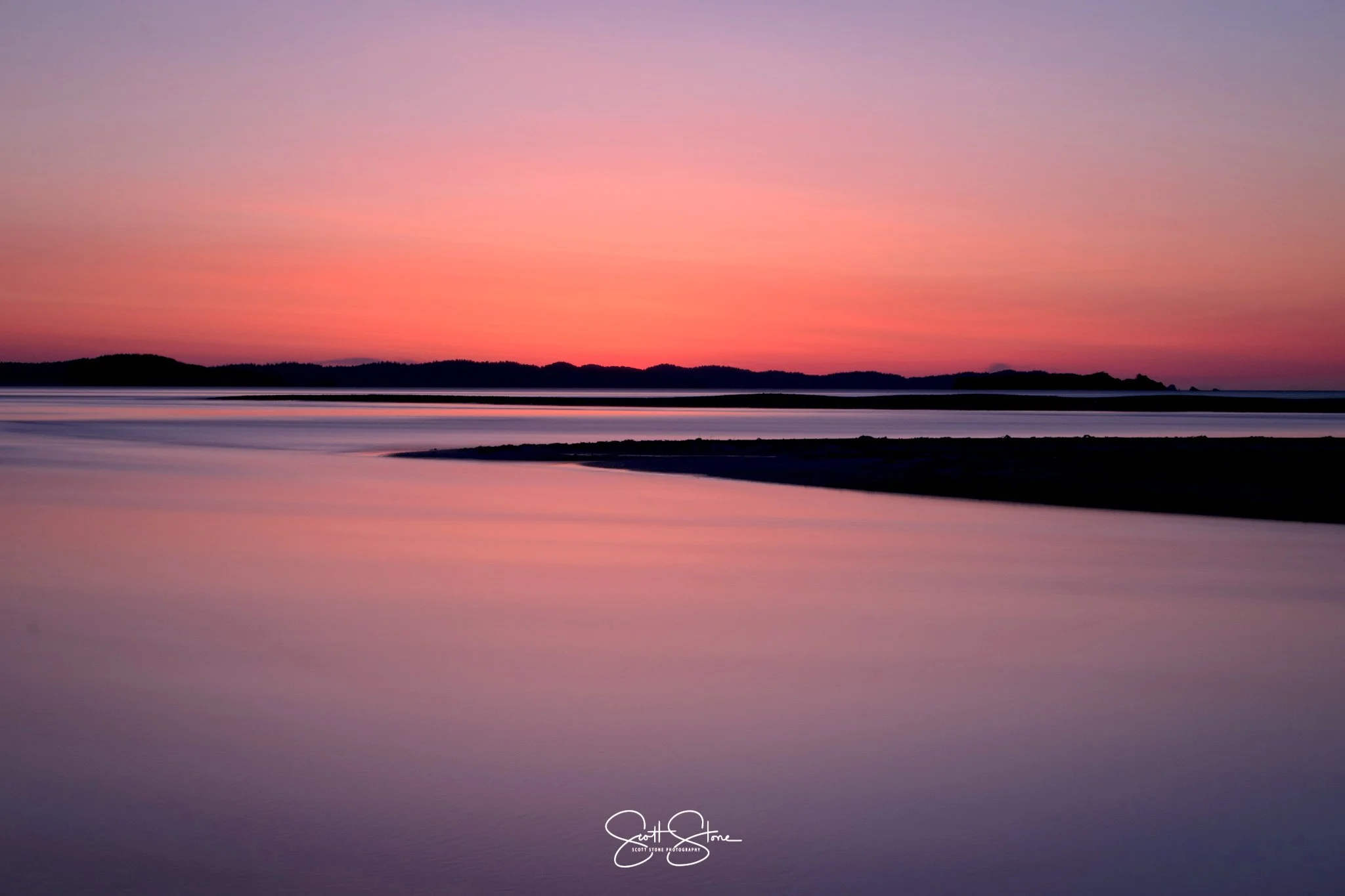 Serene sunset over calm water reflecting pink and purple hues, with silhouettes of distant land across the horizon.