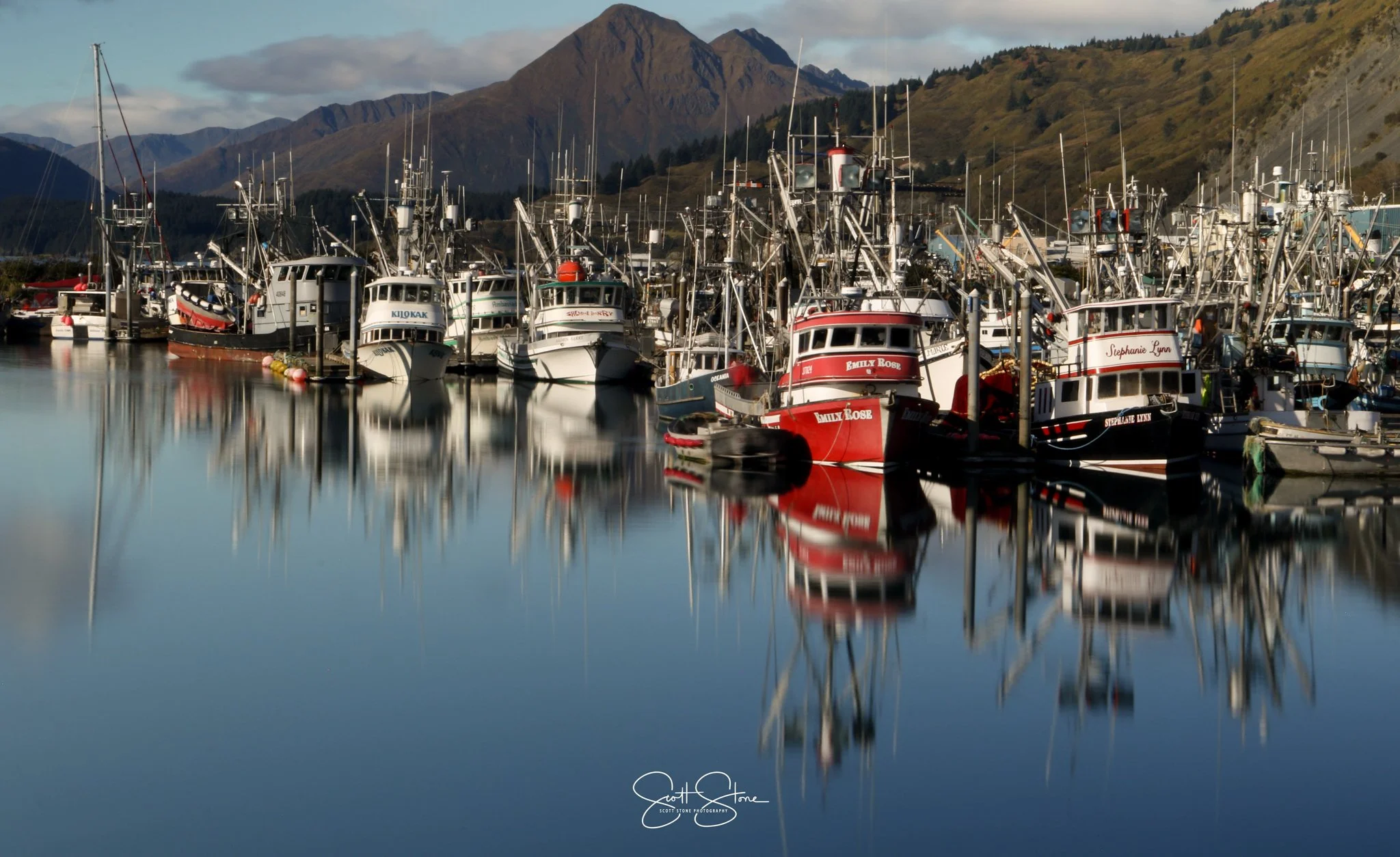 A marina filled with many fishing boats with their reflections visible in the calm water, mountains in the background, and partly cloudy sky.