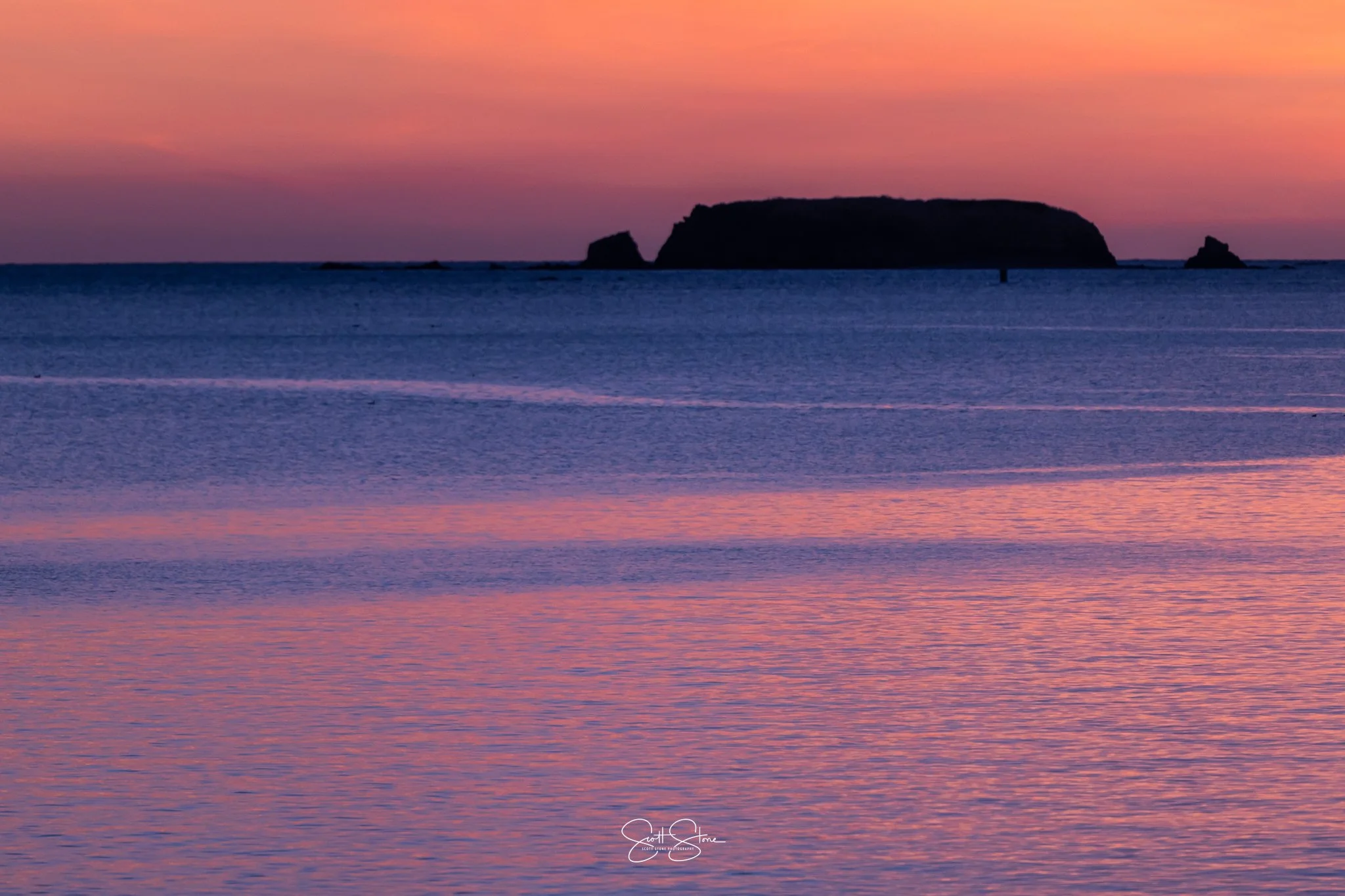 A serene ocean scene at sunset with pink, purple, and orange hues in the sky and reflections in the water, featuring a silhouette of a large rock formation or island in the distance.