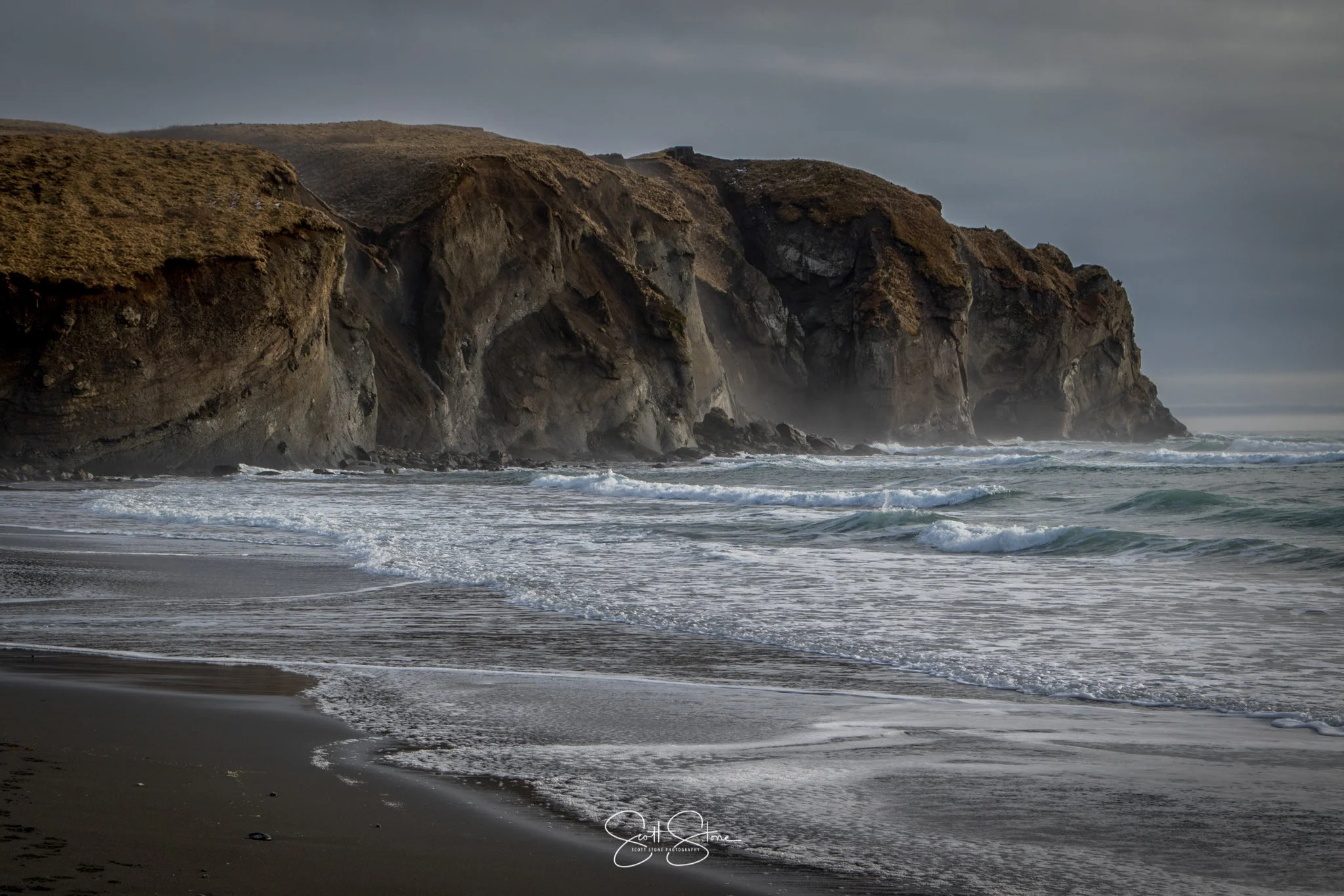 Coastal scene with dark, rocky cliffs and overcast sky, waves crashing onto black sandy beach.