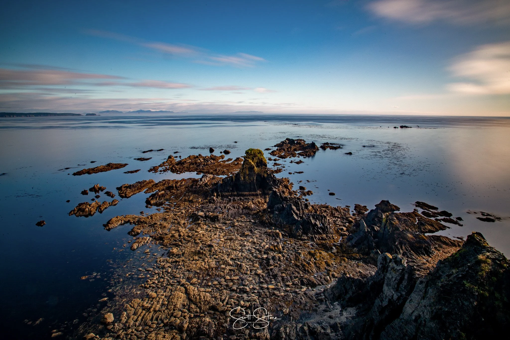 Coastal scene with rocky shoreline, calm water, and a distant horizon under a partly cloudy sky.