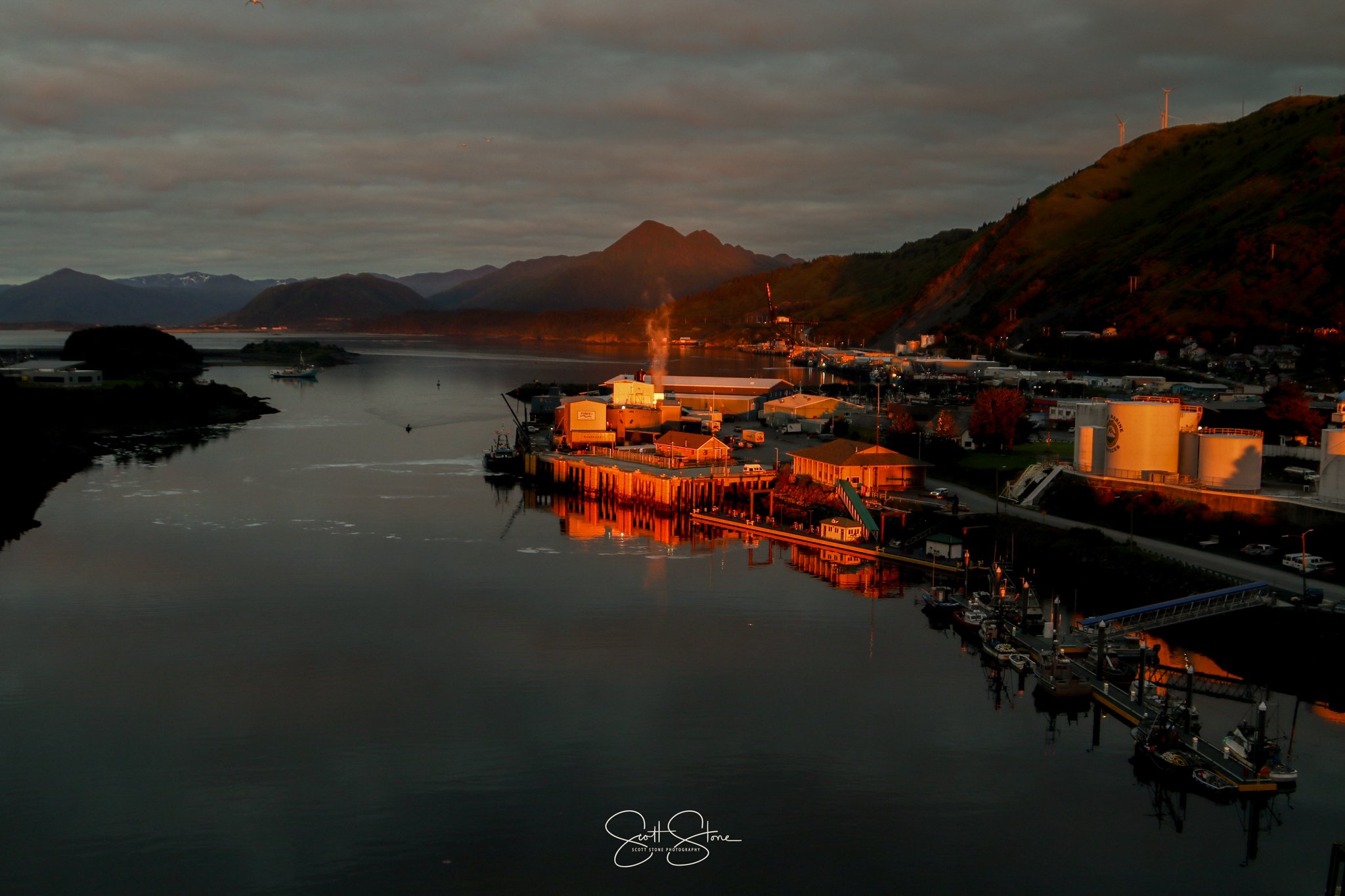 Sunset over a harbor with buildings, boats docked at piers, mountains in the background, and cloudy sky.