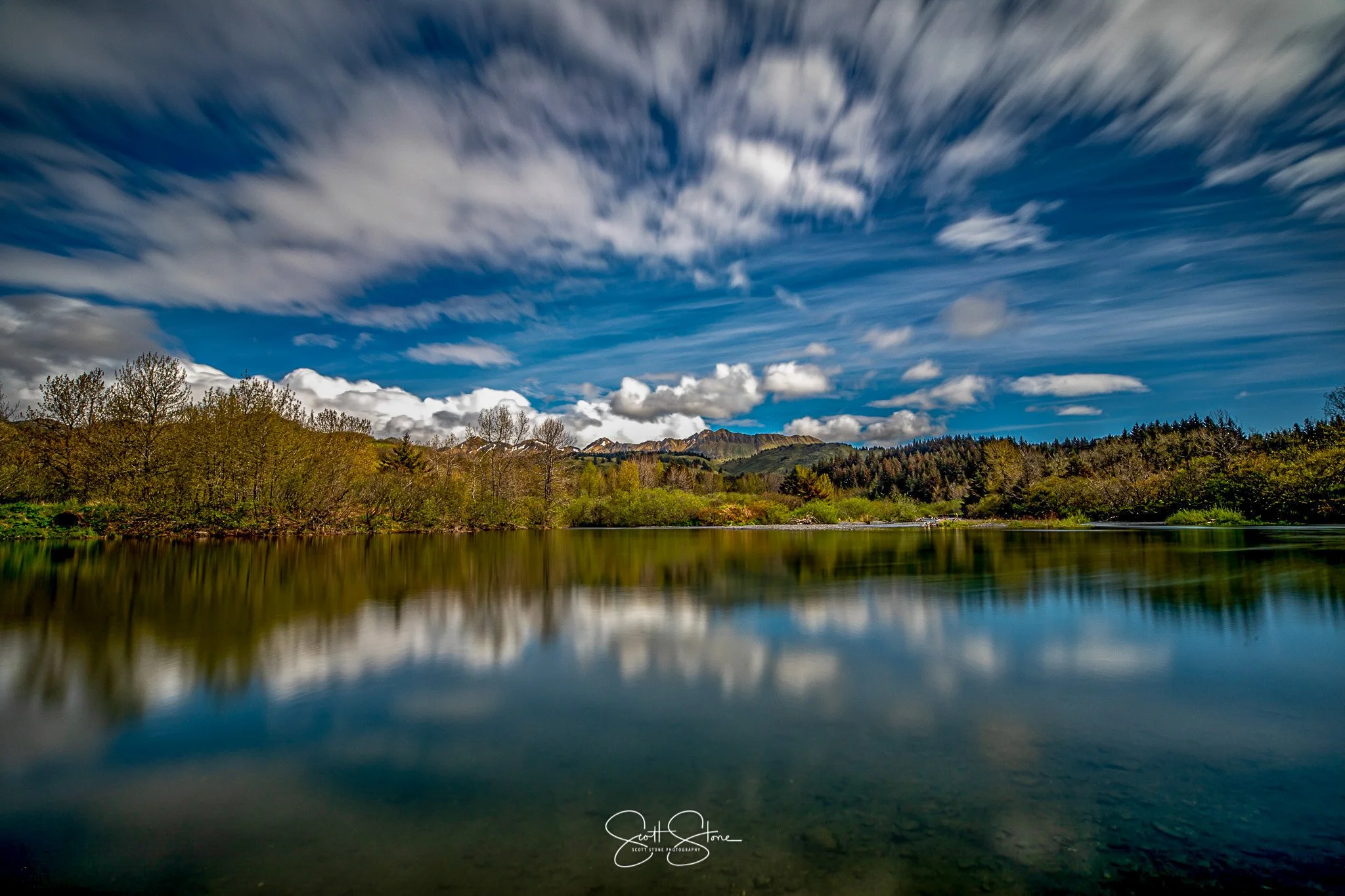 A calm lake reflecting trees and a mountain range under a partly cloudy sky.