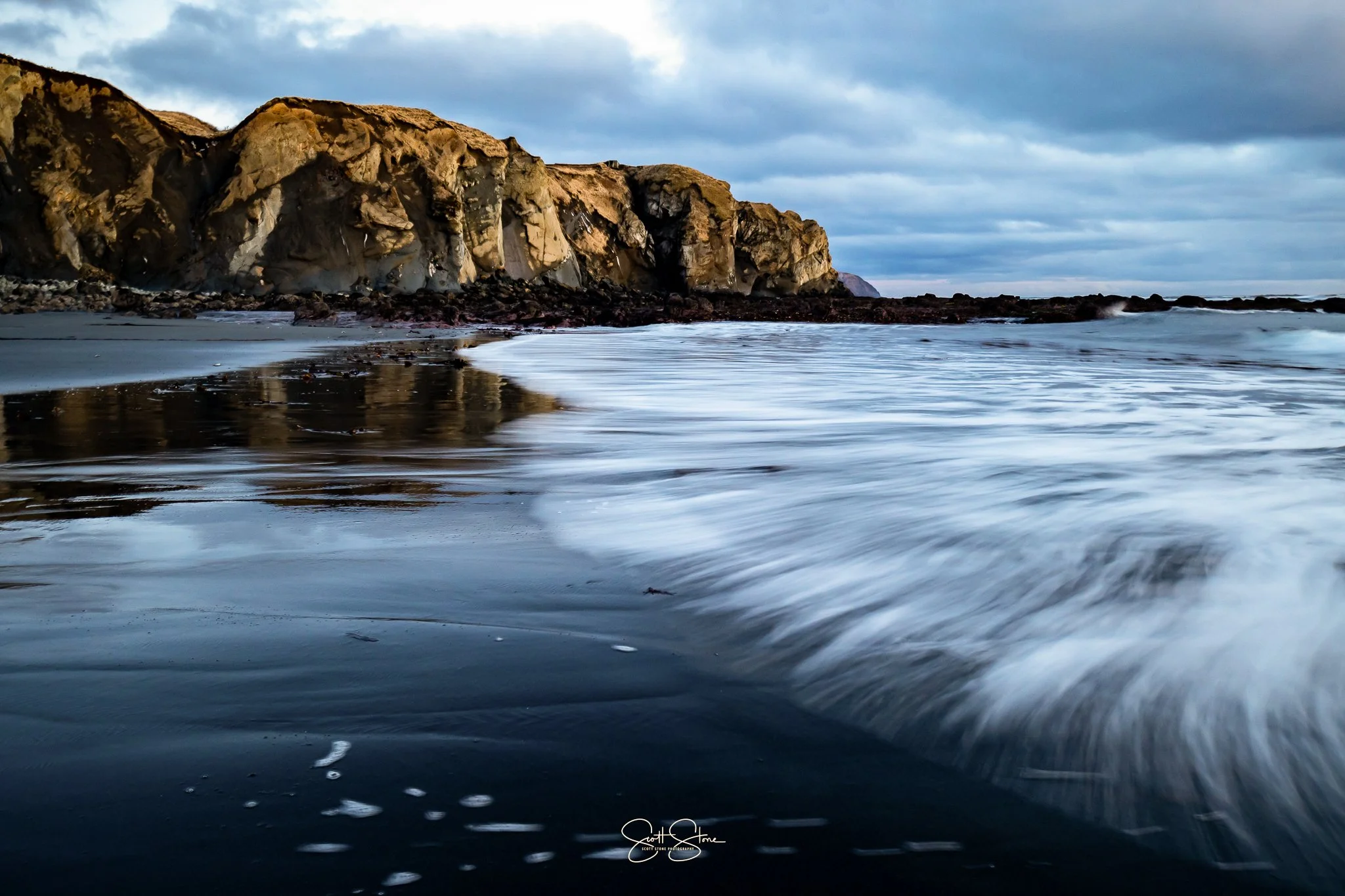 Ocean waves washing on a dark sandy beach with rocky cliffs in the background and cloudy sky overhead.