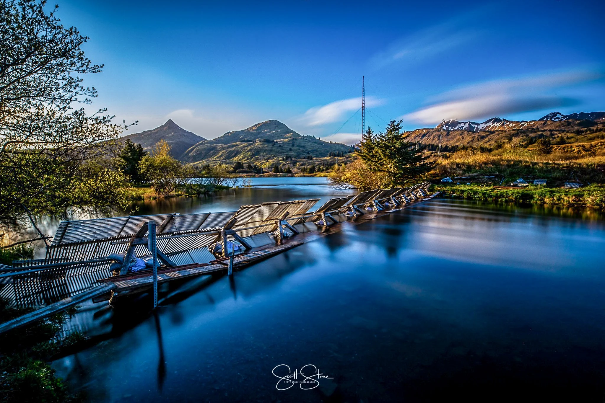 Damaged metal fencing submerged in a calm river with mountains, trees, and a blue sky in the background.