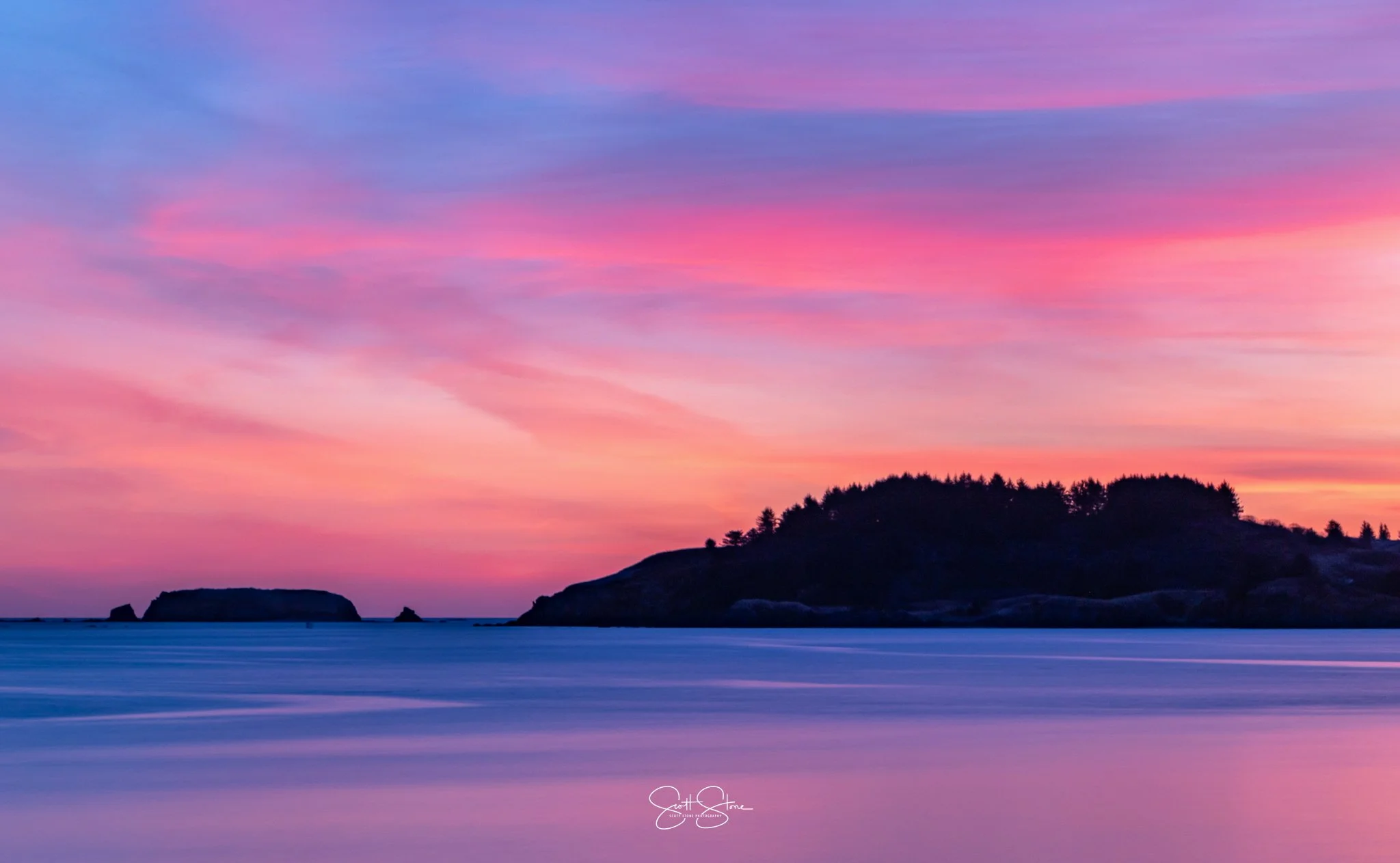 A colorful sunset over the ocean with pink, purple, and orange hues in the sky, and silhouetted land formations and an island in the distance.