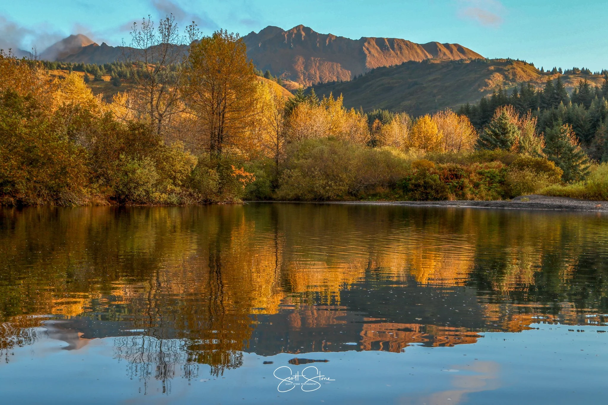A serene lake surrounded by trees with colorful autumn foliage, with mountains in the background and a clear sky above.