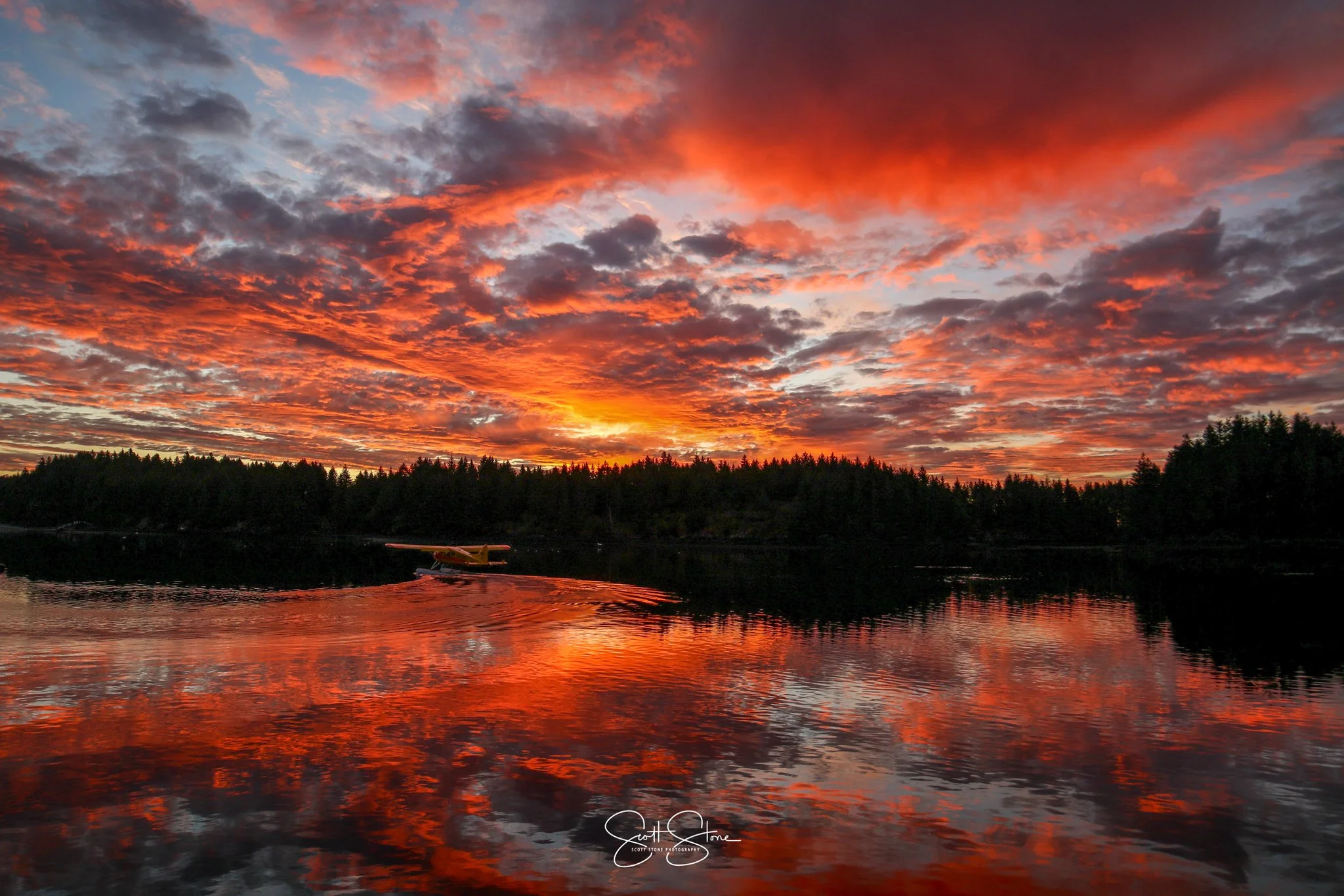 A vibrant sunset over a body of water reflects orange, red, and purple colors in the sky and on the water. Silhouettes of trees line the horizon with a small boat on the water.