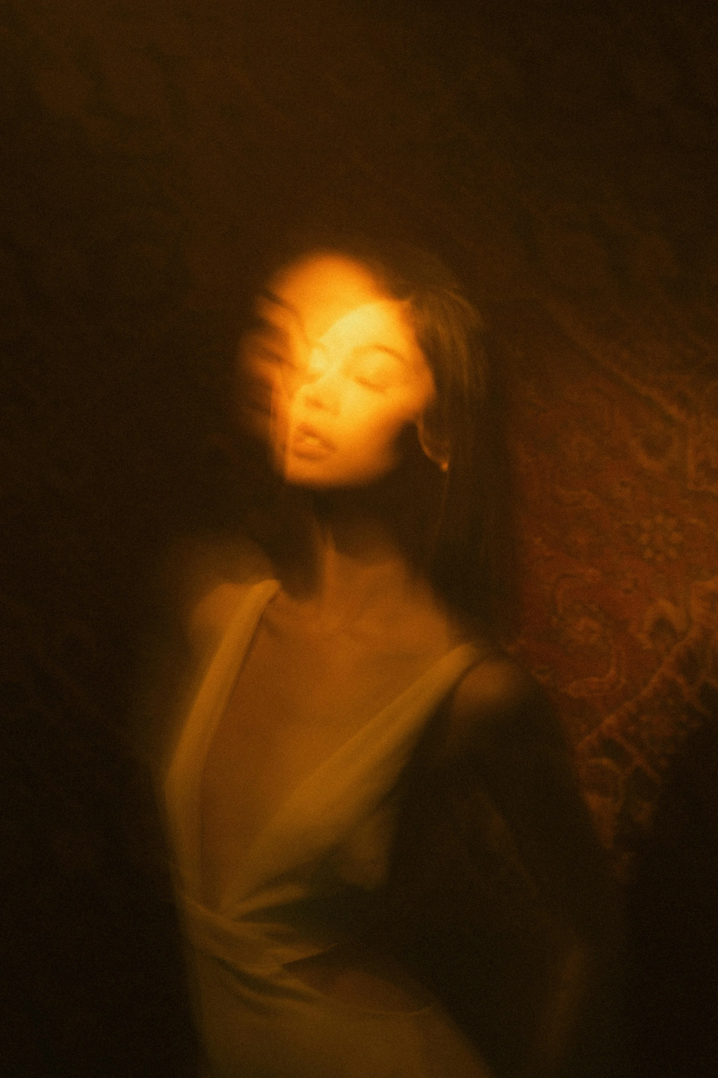 A woman with dark hair, wearing a sleeveless dress, standing against a textured wall, with a warm, blurred lighting effect creating an artistic, dreamy atmosphere.