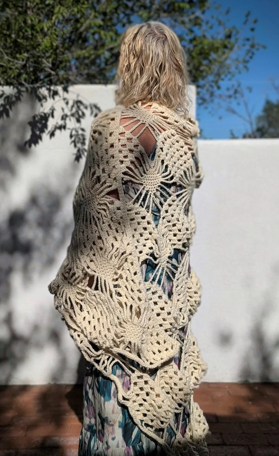 Person with blonde curly hair, wearing a large beige crochet shawl designed by Sylvia O'Regan with an intricate openwork pattern, standing outdoors on a wooden deck, with a white wall and green trees in the background under a clear blue sky.