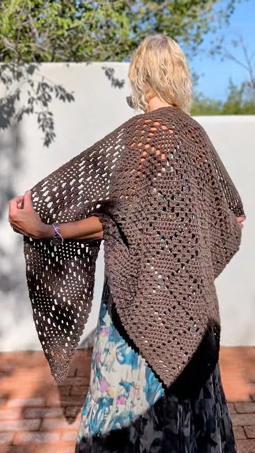 A woman with blonde hair wearing a brown crocheted shawl over a floral dress, standing outdoors on a brick patio with a white wall and greenery in the background.