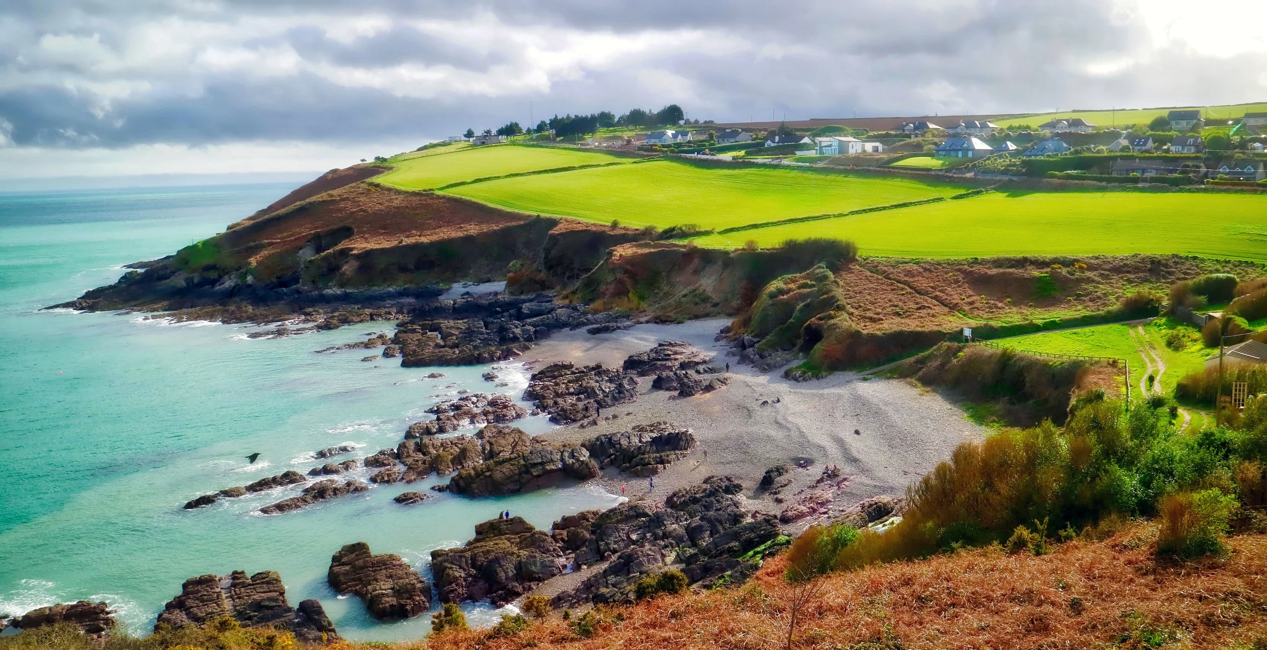 Coastal Irish landscape with rocky shoreline, sandy beach, green grassy cliffs, and houses on a hillside under a partly cloudy sky. Copyright 2026 by Pinky Brand Photography.