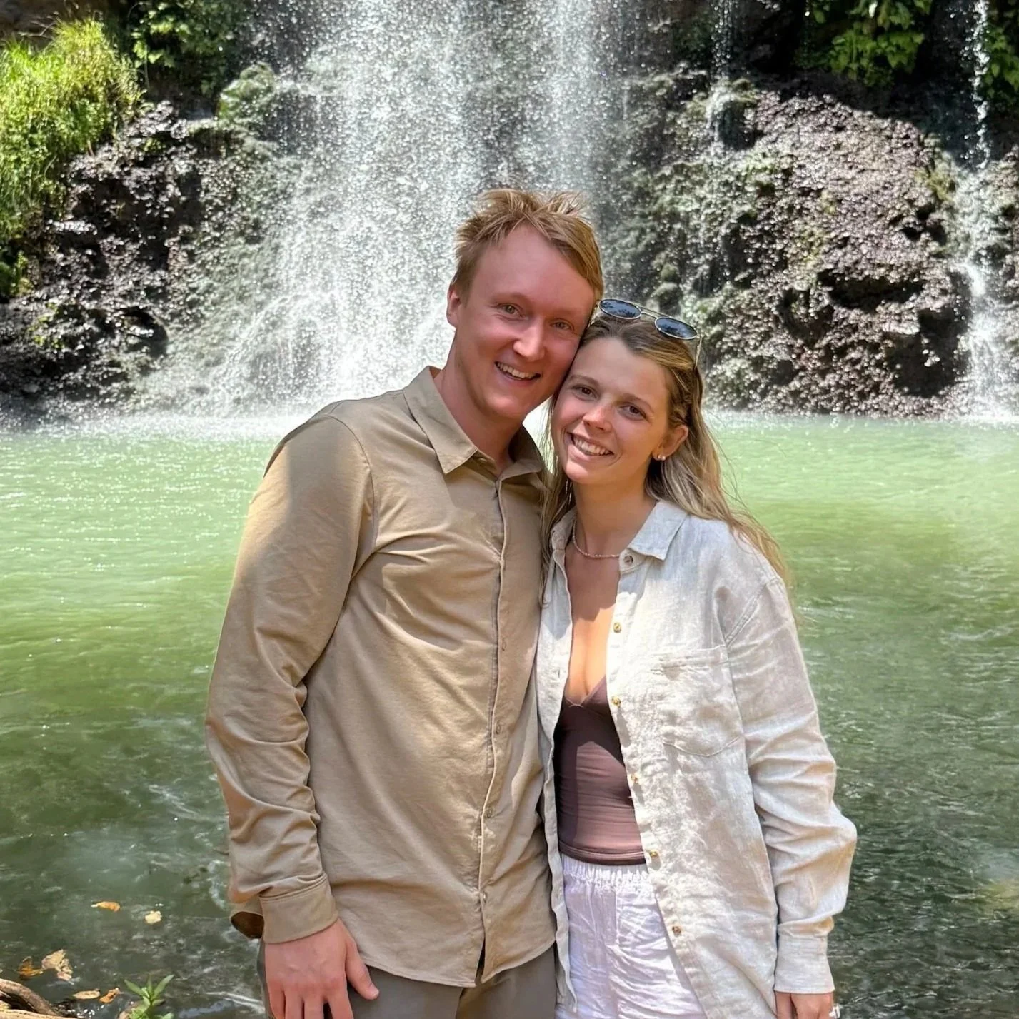 A smiling man and woman are standing close together in front of a waterfall and a green river. The woman is wearing sunglasses on her head and a light-colored jacket, while the man is in a beige shirt.