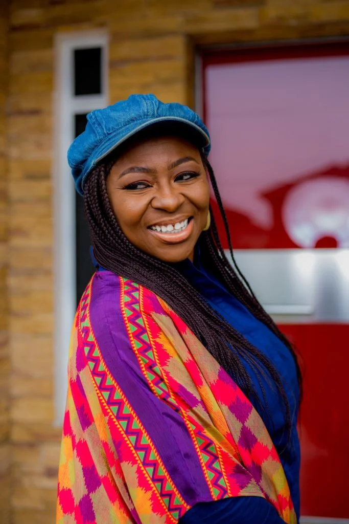 A woman with braided hair smiling, wearing a blue cap and a colorful, patterned shawl, standing in front of a building with a brick wall and a red door.