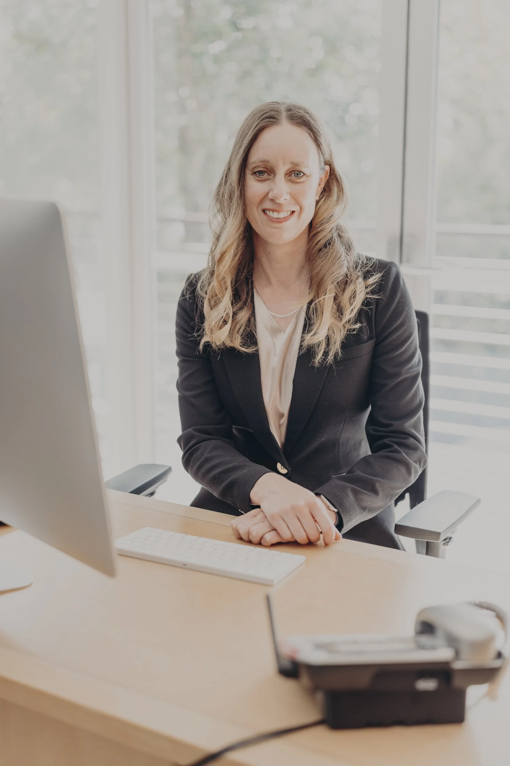 A female orthopaedic surgeon dressed in business attire sitting at a desk with a computer, and office supplies, smiling in a well-lit office with large windows.