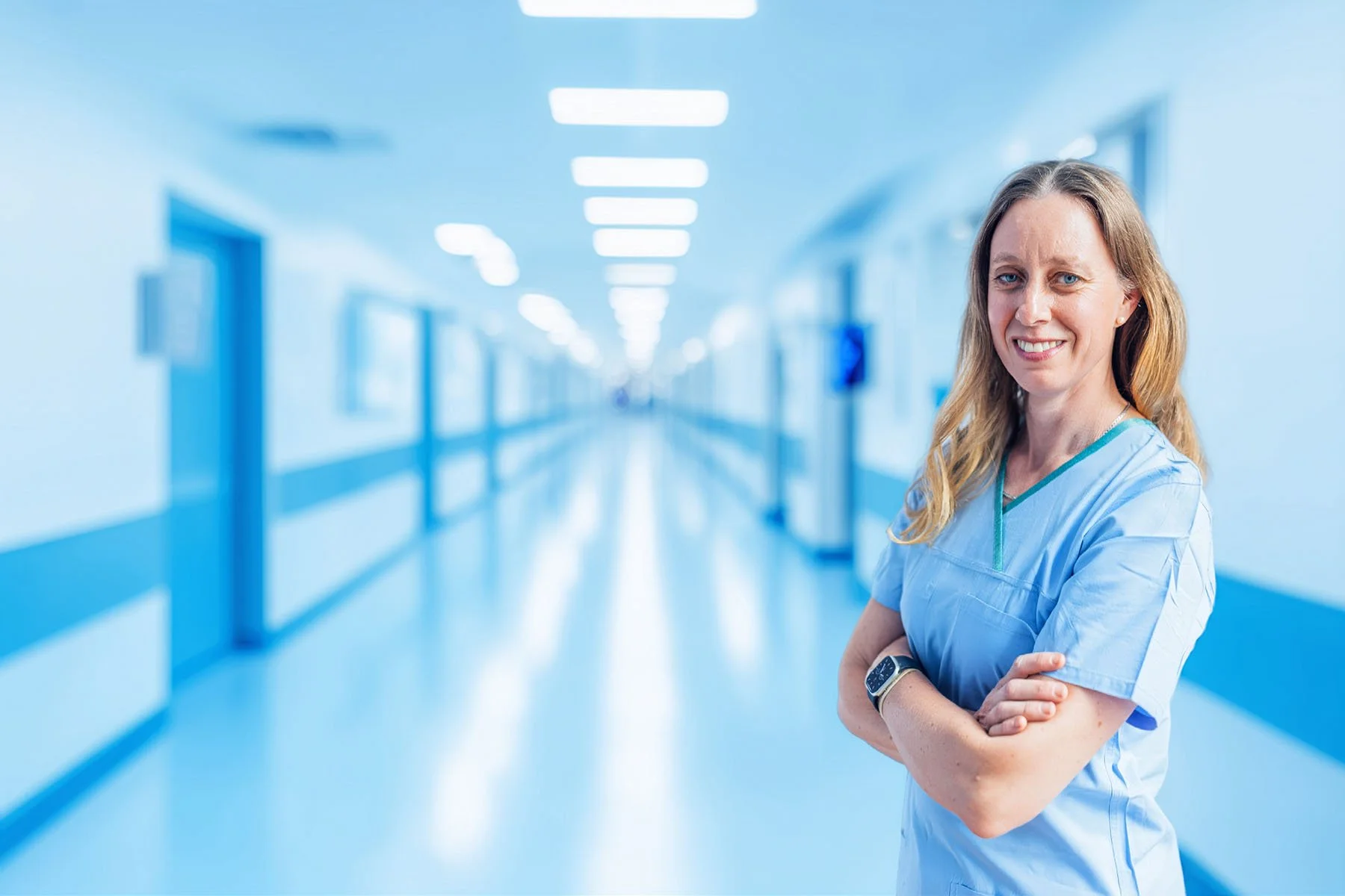 A female orthopaedic surgeon in scrubs standing in a hospital corridor, smiling with arms crossed.